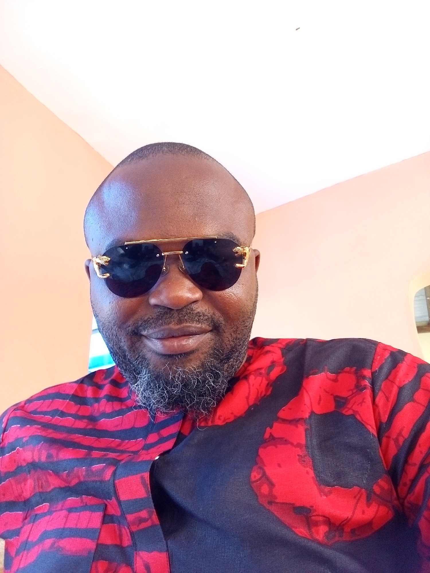 A medium-shot photograph depicts a man wearing sunglasses. He has a dark complexion, a shaved head, and a graying beard. He is dressed in a red and black patterned shirt. The background is a plain wall with a light peach color. The location in Ankpa, Nigeria is not evident in the image.