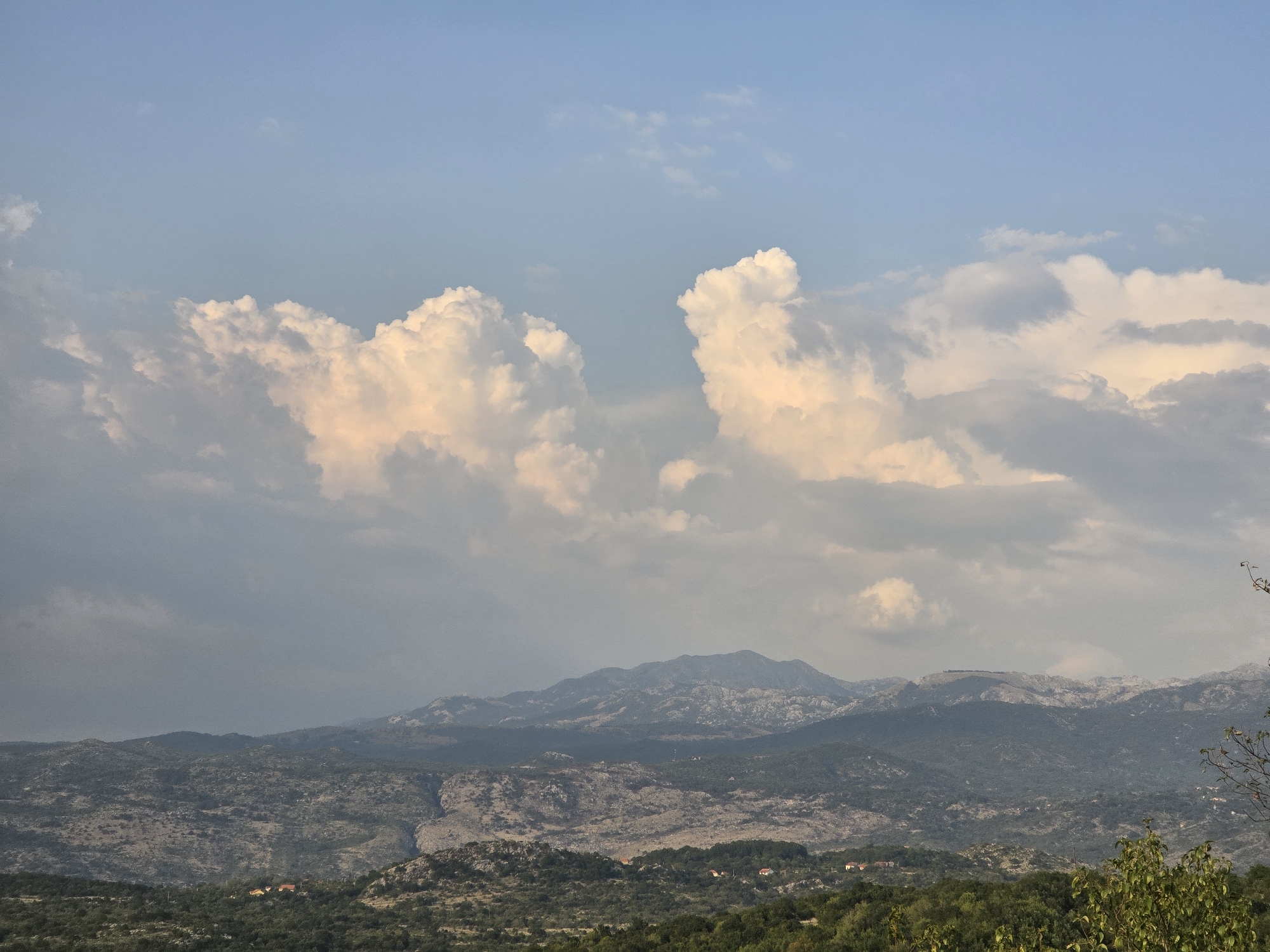 This is an eye-level, medium shot of a mountainous landscape with a cloudy sky, likely captured in the vicinity of Medun, Montenegro. The foreground is dominated by lush green trees, with a few small houses with red roofs scattered amongst them. The midground features a rugged, rocky mountain range, sparsely vegetated in some areas and densely wooded in others. A deep ravine is visible cutting through the mountains. In the background, larger, more imposing mountains rise, their peaks intermittently covered in lighter colored rock or snow. The sky is a pale blue, partially obscured by large, dramatic cumulus clouds that are illuminated with a warm, golden light, suggesting it is either sunrise or sunset. The overall impression is one of natural beauty and a remote, serene environment.