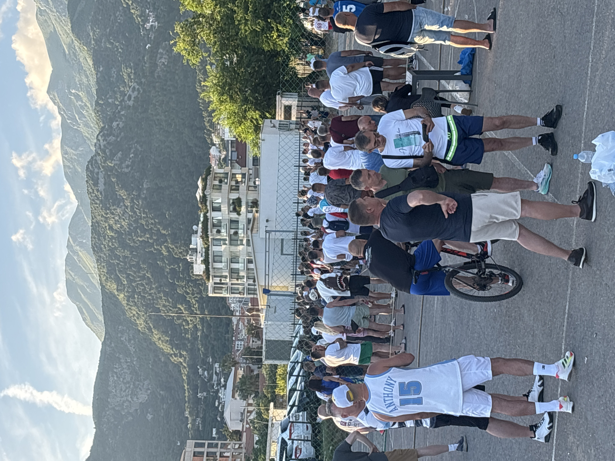 The image depicts a large outdoor gathering of people, predominantly adult men, standing on a paved surface in what appears to be a queue or a casual assembly. The scene is set against a backdrop of majestic, tree-covered mountains under a clear, bright sky with some scattered clouds, characteristic of Budva, Montenegro. Modern multi-story buildings, likely residential or hotels, are visible between the crowd and the mountains, with smaller structures nestled into the hillsides.
Most individuals are dressed casually in t-shirts, shorts, and sneakers. In the foreground, one person wears a white basketball jersey with "ANTHONY" and the number "15" clearly visible on the back. Another person wears a white t-shirt with partial text "NEW". A black bicycle is parked among the crowd, and a plastic water bottle and bag are seen on the ground. A metal fence separates different sections of the area.
The time of day appears to be late afternoon or early evening, indicated by the warm, directional sunlight casting long shadows across the ground and illuminating the mountainsides. The atmosphere seems relaxed, with people standing and waiting, though no strong emotions are discernible. The event could be a public viewing, a fan gathering, or a local social event given the casual attire and large attendance.