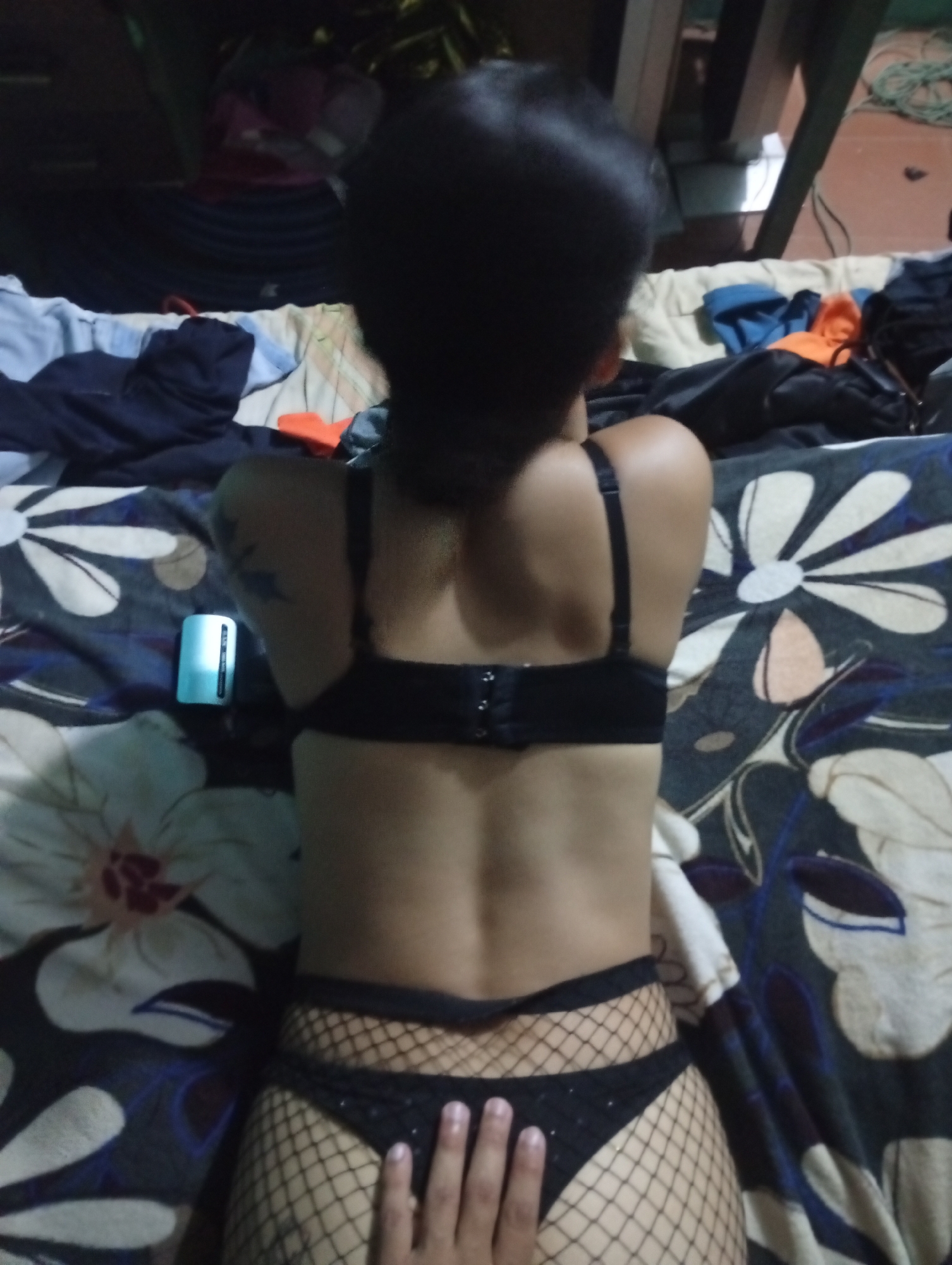 A person is lying on their stomach on a bed with a floral patterned bedspread. They are wearing a black bra and black fishnet stockings with a black thong underneath. The person's head is turned away from the camera. A hand is touching their lower back. There is a small, light blue object to the left of the person. Other objects, including clothing, are visible on the bed and in the background. It's not possible to confirm the location as Altagracia, Venezuela, from the image alone.