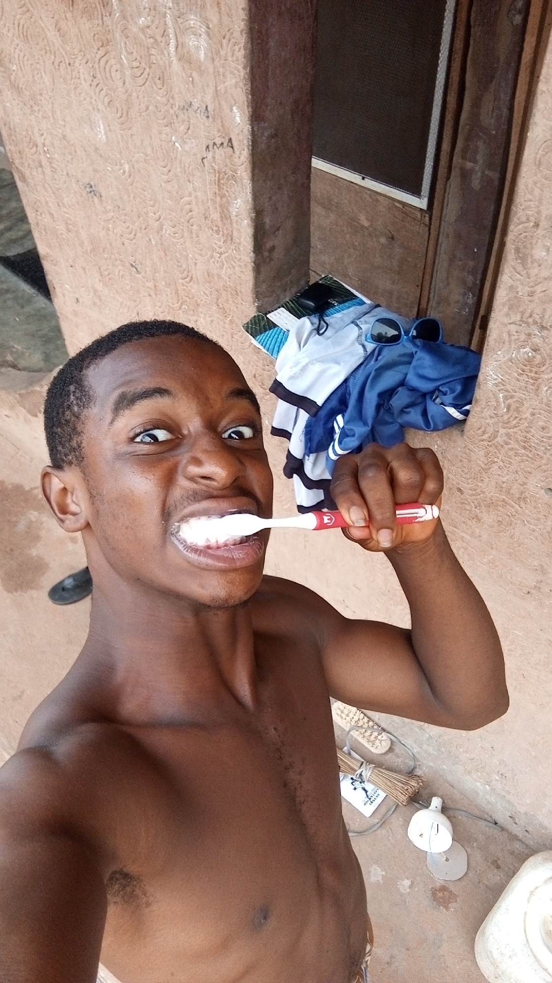 A shirtless, dark-skinned man with short hair is brushing his teeth with a red and white toothbrush. His eyes are wide and he is grinning. He is in an outdoor setting near what appears to be a wall. Behind him are a pile of clothes, sunglasses, and a window. On the ground are a broom and a container. The exact location within Onitsha, Nigeria, cannot be determined with the provided image and information.
