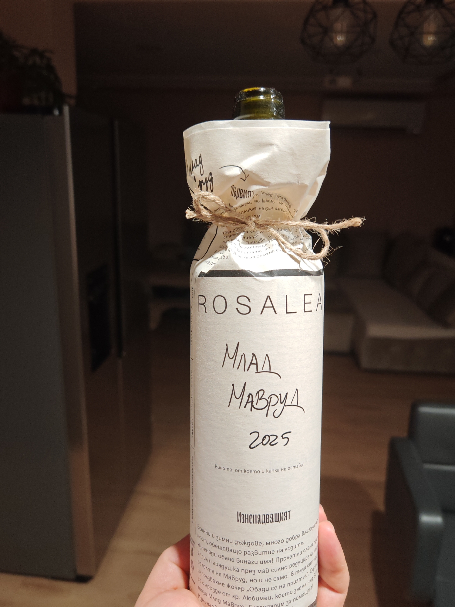 The image shows a hand holding a wine bottle with a rustic, handmade label. The label has handwritten text in Bulgarian. The bottle is wrapped with a piece of parchment-like paper tied with twine. The text on the label mentions "МЛАД МАВРУД 2025", which translates to "Young Mavrud 2025". Mavrud is a local red wine grape variety from Bulgaria, and Plovdiv is its region of origin. The background appears to be an indoor setting, possibly a home, with modern furniture and lighting visible.