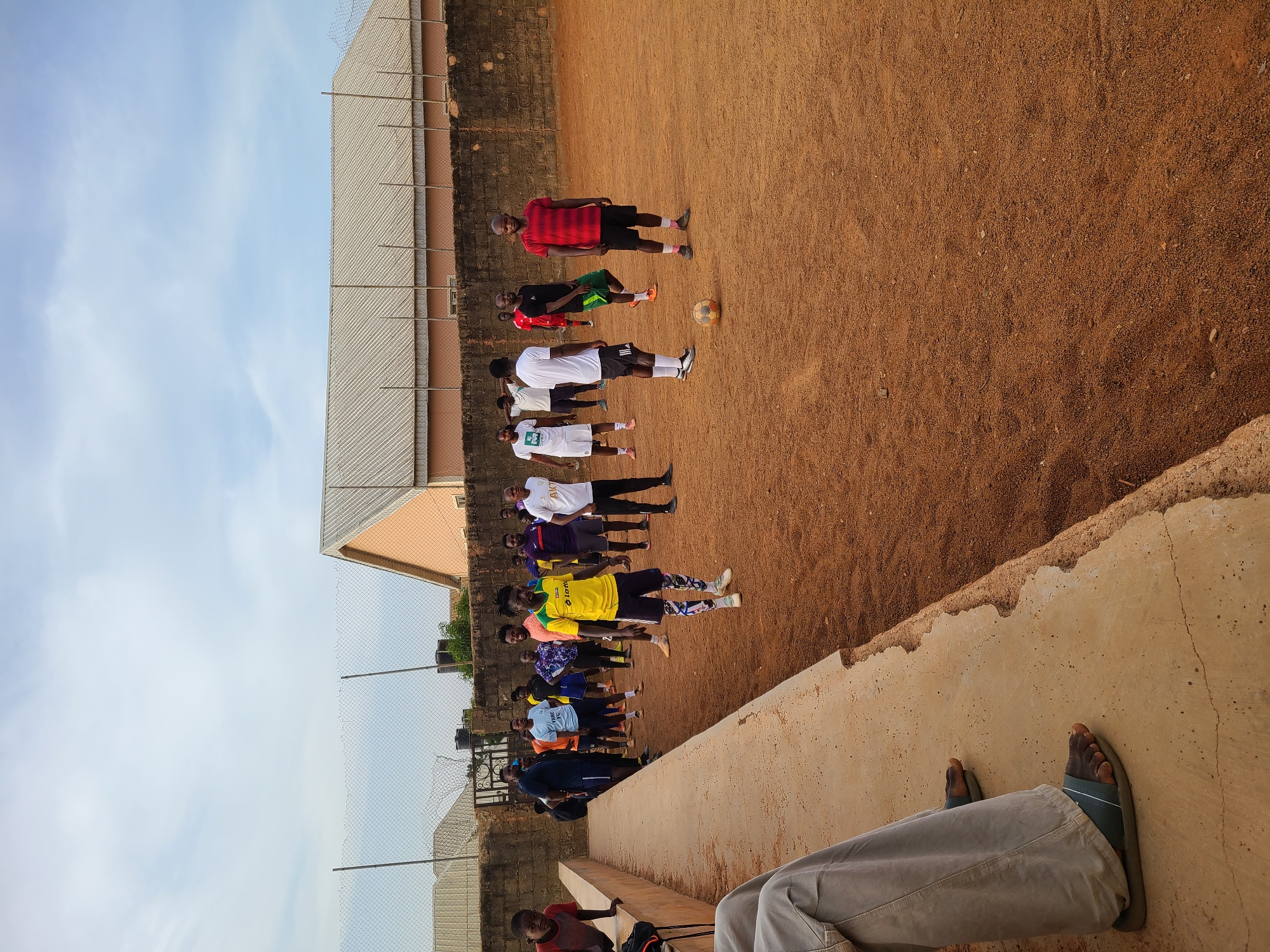 A soccer practice with about 13 players is taking place on a red clay field next to a concrete ledge and fence. Several of the players are standing with their backs to the camera, with a soccer ball visible near the center of the field. An individual wearing pants and sandals is sitting on the ledge, presumably watching the practice. The location is in an urban environment, with buildings visible behind the fence.