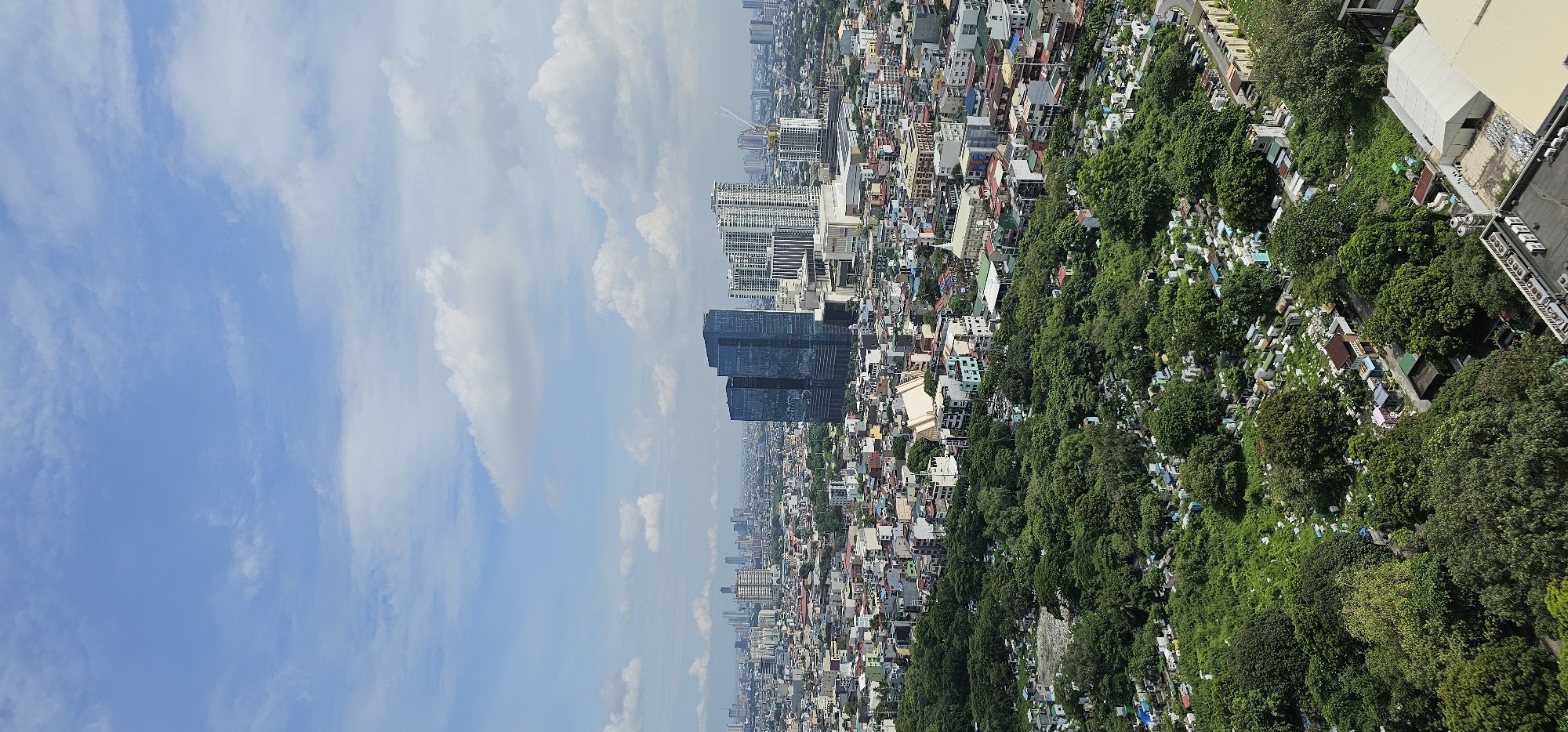 An aerial shot captures a dense urban landscape with a prominent cemetery in the foreground. Lush green trees and numerous white tombstones fill the lower portion of the frame, interspersed with some smaller structures. Beyond the cemetery, a sprawling city unfolds with a mix of residential buildings, smaller commercial establishments, and significant modern skyscrapers. Two striking, dark glass skyscrapers stand out in the mid-ground, flanked by a cluster of lighter-colored, multi-story buildings. The background reveals a hazy, distant skyline with more buildings and a pale blue sky with scattered white clouds. The scene appears to be in Makati, Philippines, characterized by its dense urban development and the presence of both older structures and contemporary high-rises. There are no discernible people or immediate actions in this static, observational view.