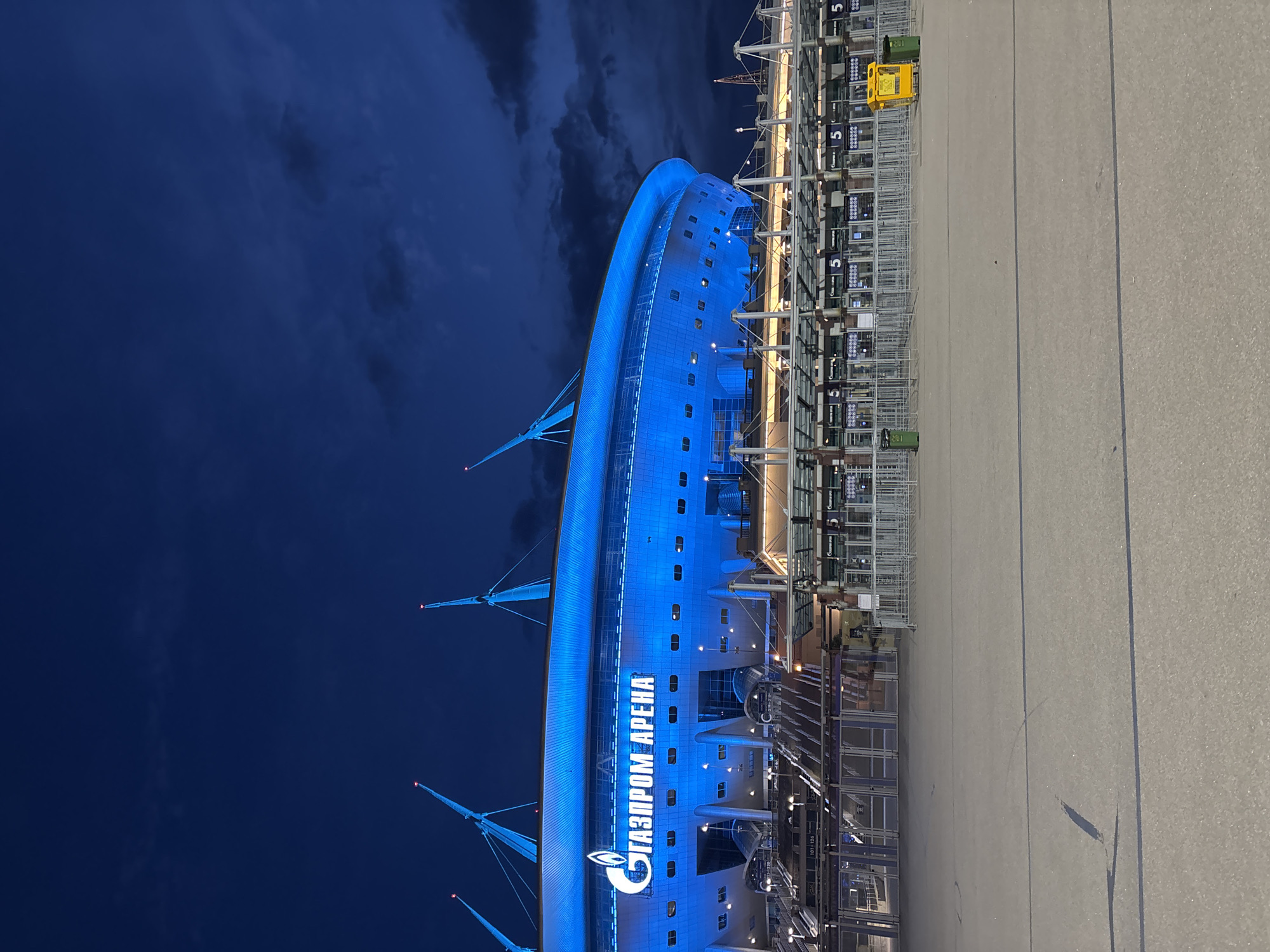 The image captures the Gazprom Arena in Saint Petersburg, Russia, at night. The impressive stadium building dominates the left side of the frame, illuminated in a bright, striking blue light against a dark blue, cloudy sky. The architecture features a smooth, curved facade with numerous small, dark windows, and several tall, slender spires or masts extending upwards from the structure, tipped with small red lights.
Prominently displayed on the side of the building is the name "ГАЗПРОМ АРЕНА" (GAZPROM ARENA) in large, glowing white Cyrillic letters, accompanied by the Gazprom logo.
To the right of the arena, there is a wide, empty paved area, likely a pedestrian plaza or concourse. Along the edge of this area, closer to the stadium, a series of metal barriers and turnstiles or entry gates are visible, suggesting an entrance point. Several signs with the number "5" are attached to these structures, possibly indicating gate numbers. A yellow cart and a green waste bin are also present near these gates. The ground is a uniform light grey, with a few faint lines and marks.
The scene is devoid of people, giving it a quiet and somewhat imposing atmosphere despite the vibrant lighting. The dark sky with visible clouds confirms it is nighttime, possibly after an event or before one starts, or simply showcasing the illuminated building. The blue lighting of the arena stands out starkly against the dim surroundings.