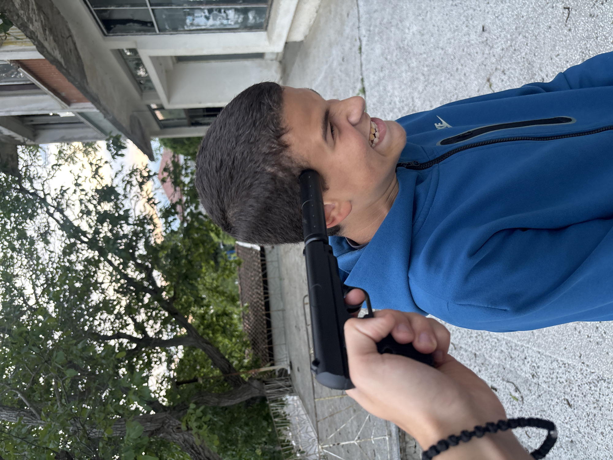A person is holding a black handgun to the head of a young man who is looking to the side and smiling. The setting appears to be outdoors, in front of a building and trees. Based on background clues like the building style and foliage, it's consistent with a setting in Goritsa, Bulgaria, or similar region.