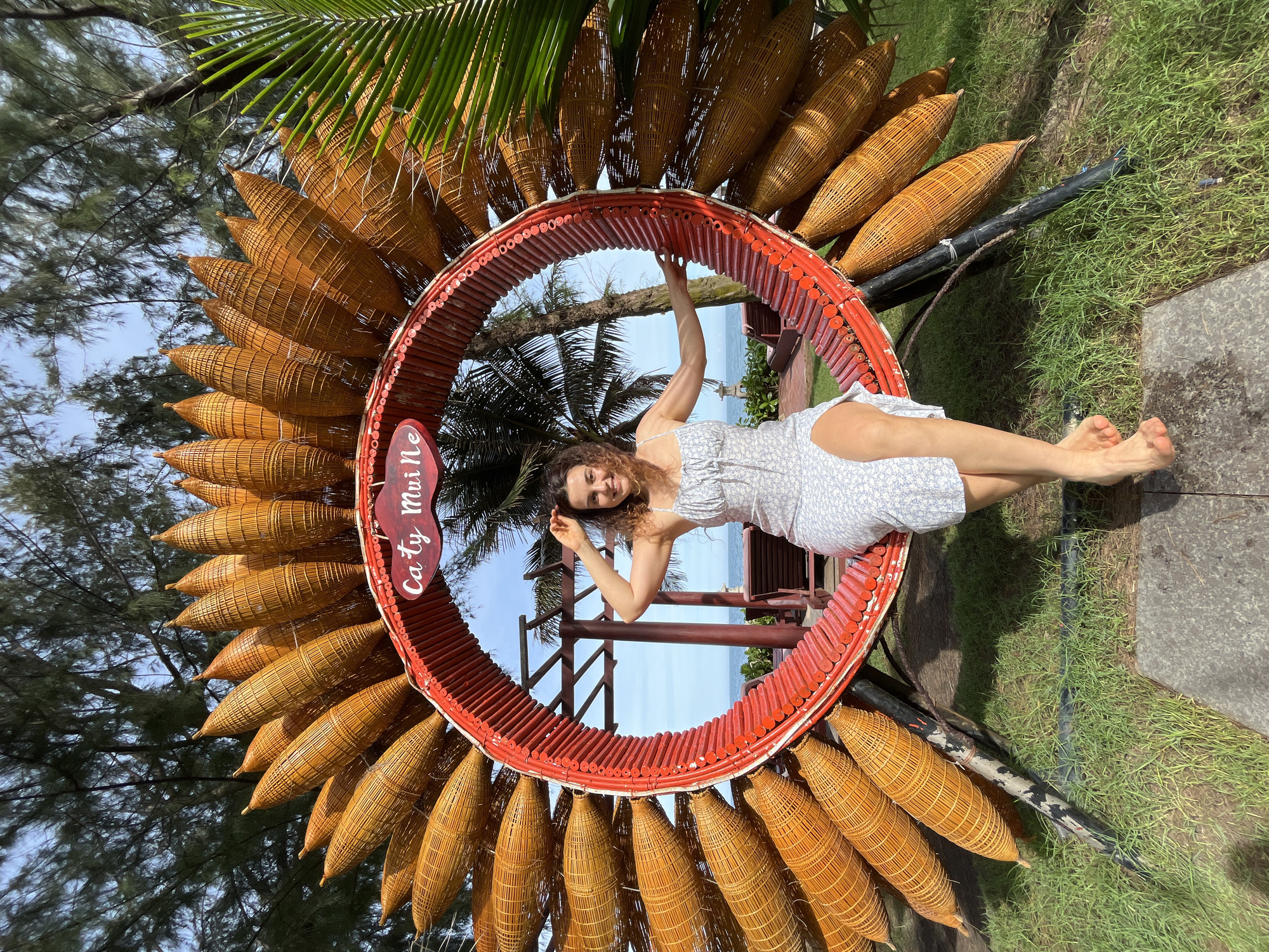 A woman in a light blue floral dress is seated on a circular, sun-shaped bamboo and red wood sculpture in Mũi Né, Vietnam. The sculpture, decorated with numerous woven bamboo pods resembling petals, frames a view of the ocean and sky. A red sign within the sculpture reads "Ca ty Mui Ne." The woman is barefoot, with her left arm resting on the sculpture and her right hand raised to her hair. The foreground features green grass and a stone path. In the background, palm trees and the ocean are visible under a bright sky.