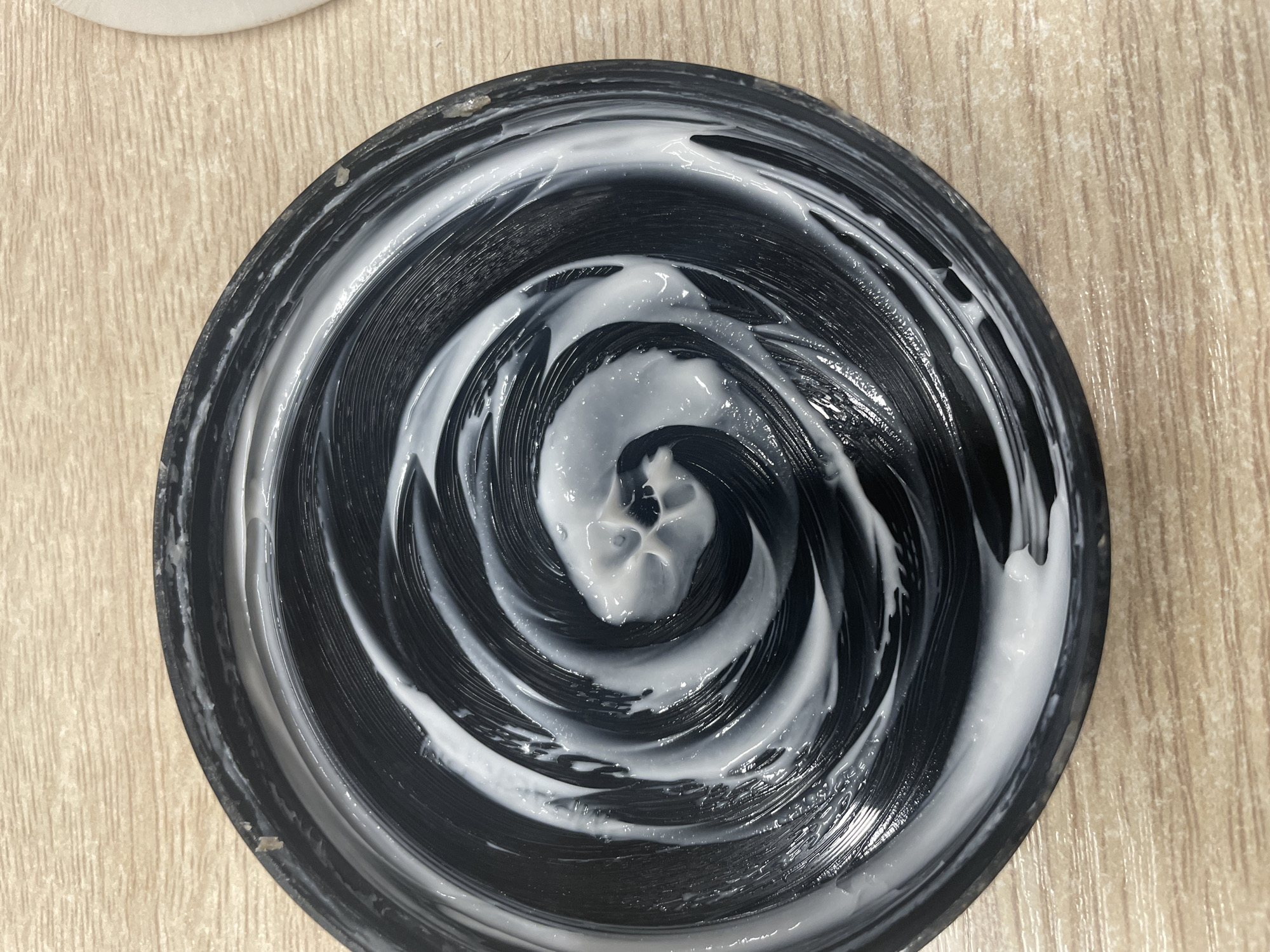 An overhead view of a black jar lid, with white cream swirled in a spiral pattern on its surface. The lid is resting on a light brown, wood-grain surface. The scene is indoors. This image is not relevant to the city of Терволово, Russia.