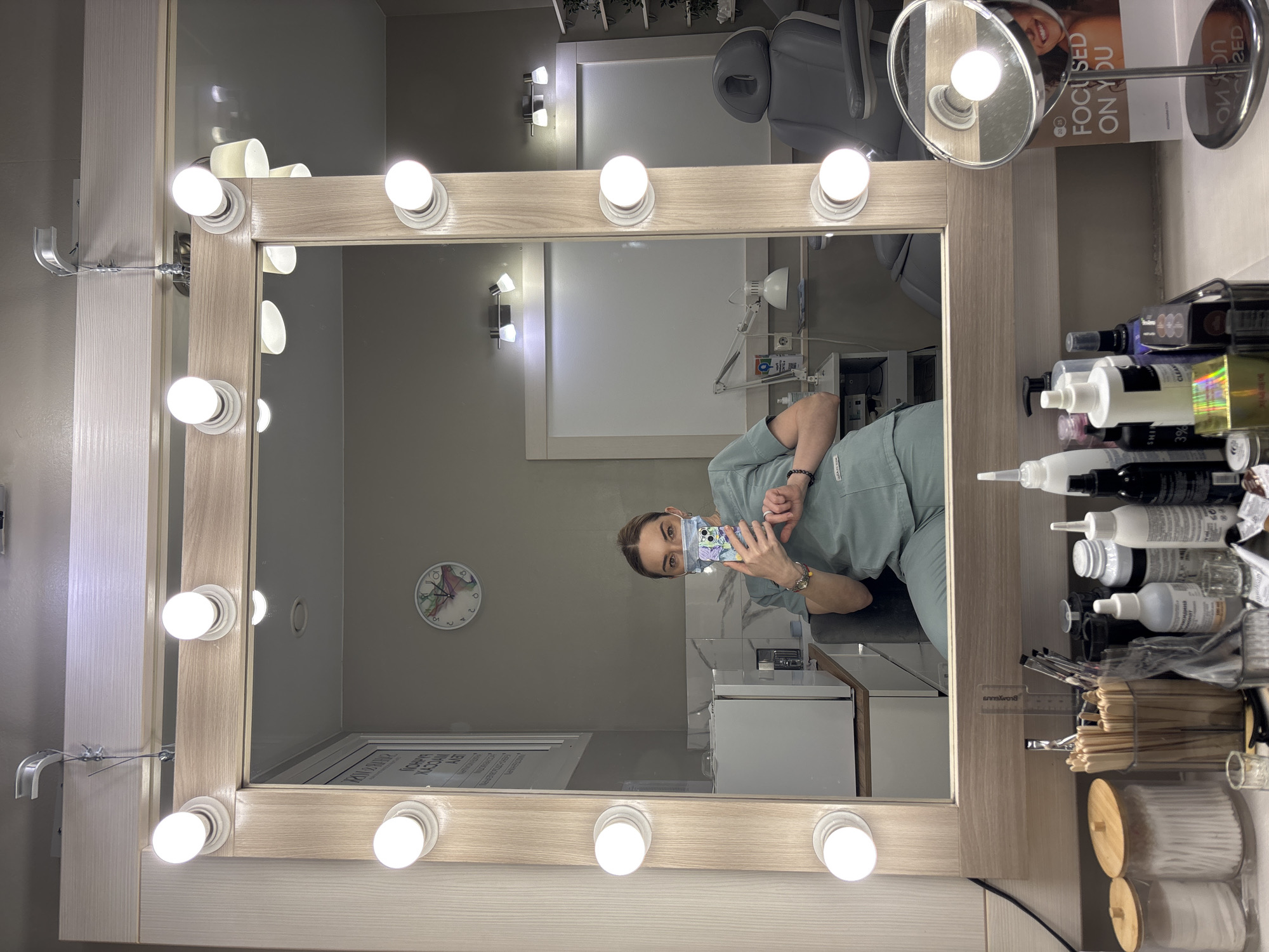 A mirror lit with vanity lights reflects a woman wearing a light green scrub suit and a face mask, holding a phone as if taking a picture. The room appears to be a medical or cosmetic treatment space, with a patient chair, a white cabinet, and various bottles and supplies on a counter to the right. A clock is visible on the wall, showing approximately 11:50. There's signage and various grooming tools on the counter near the mirror.