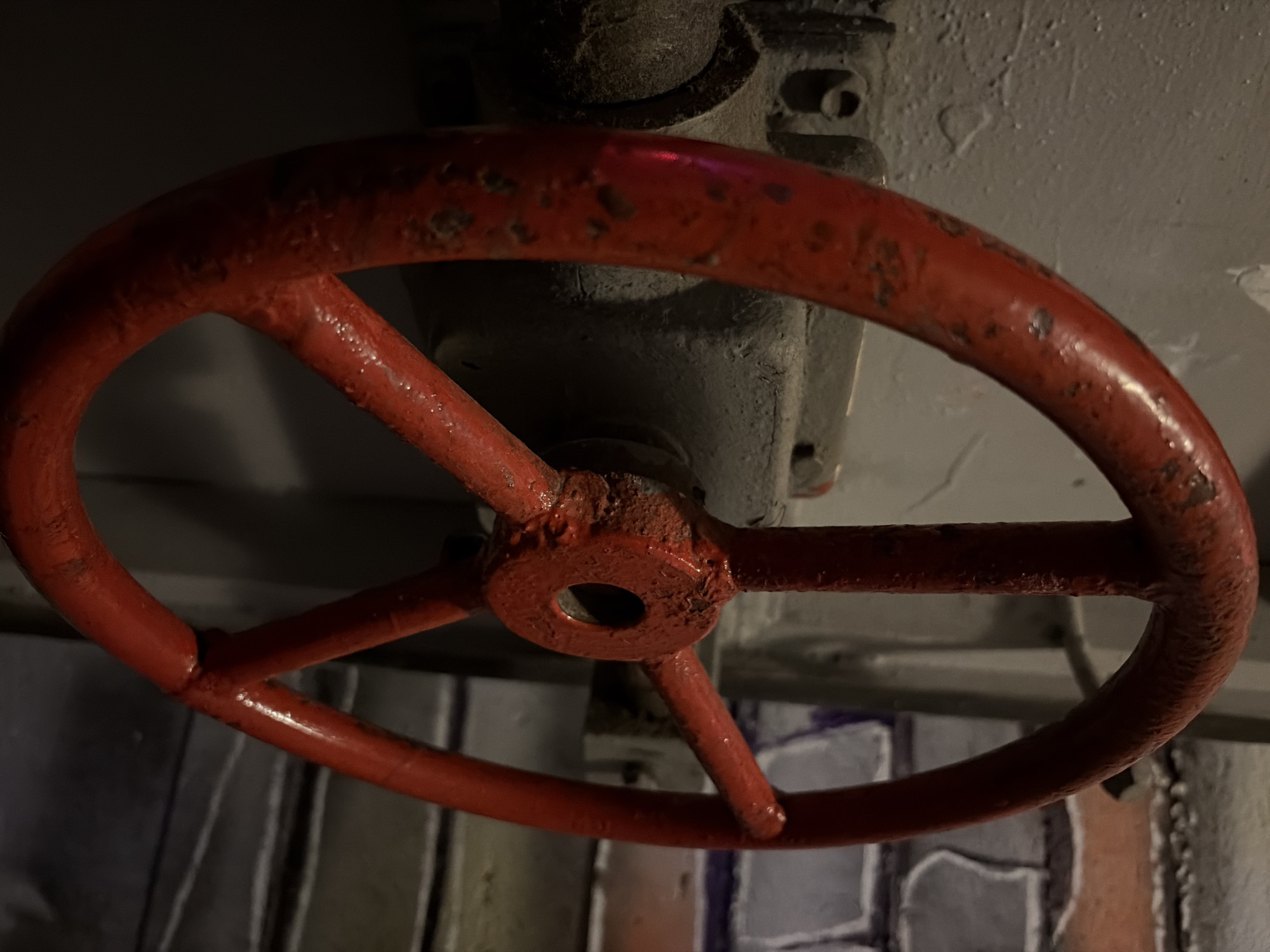 The image shows a close-up of a large, red valve wheel mounted on a gray mechanical apparatus, likely a valve or pipe control system. The wheel has three spokes and shows signs of wear and rust, with a textured surface. The valve apparatus behind it is gray, with some visible bolts or fasteners. The background is a mottled gray and white wall. The image does not provide enough information to place it in Moscow, Russia, or any other specific location. It focuses on the mechanical details of the valve and its immediate environment.