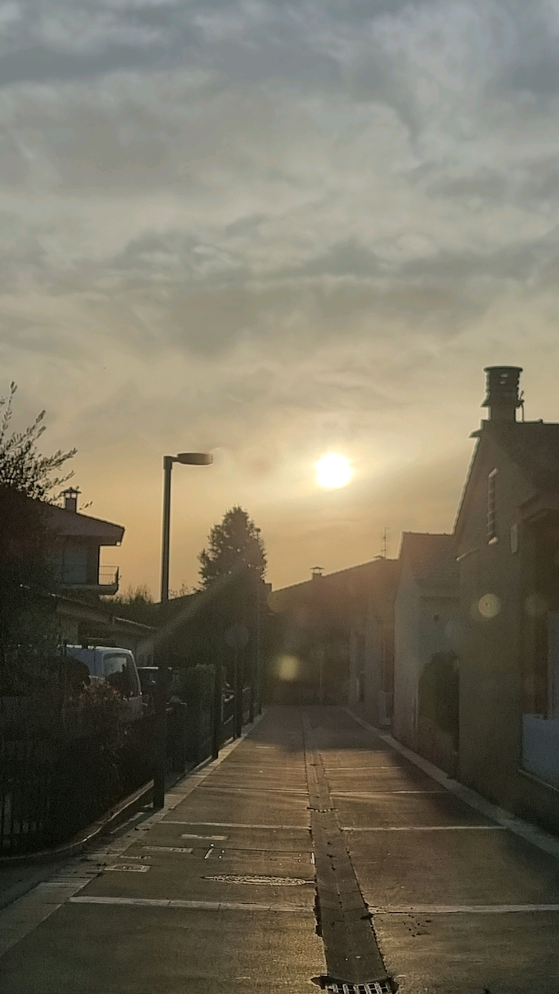 The image depicts a street scene in Sant Joan Les Fonts, Spain, during sunset or sunrise. The sun, a bright orb, is positioned in the upper right quadrant of the sky, casting a warm, hazy glow over the environment. Clouds are present in the sky, appearing somewhat diffused due to the light.
In the foreground, a paved street runs down the center of the image, reflecting the ambient light. Markings on the street, possibly lane dividers or pedestrian crossings, are visible. A dark, raised channel or drain runs along the center of the street. On either side of the street, residential buildings with tiled roofs are visible, partially silhouetted by the low sun. Some trees and vegetation line the street, adding greenery to the scene. A street lamp with a circular fixture stands tall on the left side of the street, its pole a dark vertical element against the sky. A white van is parked on the left side of the street, near some of the buildings. The overall impression is one of a quiet street at the end or beginning of the day.
