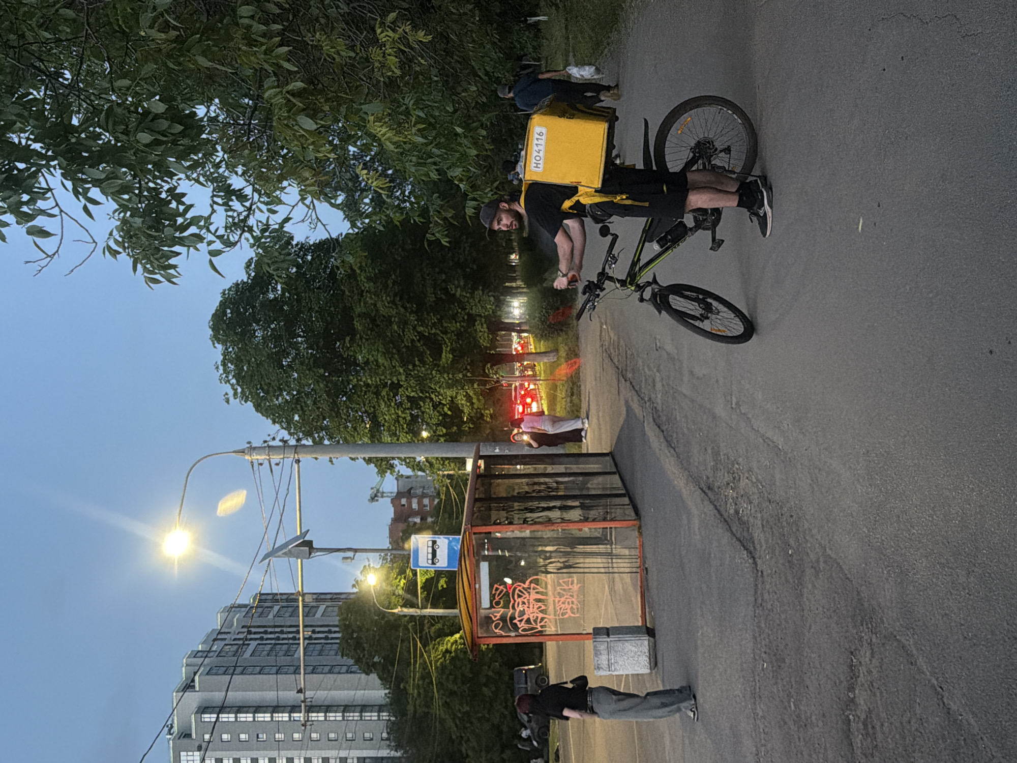 The scene depicts a street in Rostov-on-Don, Russia, at dusk. A delivery cyclist, wearing a yellow delivery backpack labeled "H04116," is standing next to his bicycle on a paved road. Further down the road is a bus stop shelter covered in graffiti. In the background, there are residential buildings and trees. A street lamp illuminates the bus stop area. Several people are present near the bus stop and further along the road, although they are not clearly identifiable. The overall impression is of a typical urban environment transitioning from day to night.