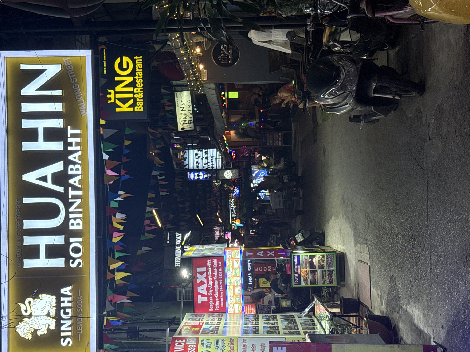 The image depicts a nighttime scene on a street in Hua Hin, Thailand, likely in the area of Soi Bintabaht, known for its vibrant nightlife and tourist services. A large, illuminated sign at the top reads "HUAHIN SOI BINTABAHT WALKING STREET," with the Singha beer logo to its left. Below this, strings of colorful triangular flags are strung across the street, creating a festive atmosphere.
Several businesses are visible. To the left, a "TAXI" sign is prominently displayed, along with contact information and service details for airport and inter-city travel. Nearby, a "Tourist Information" booth is set up with brochures. Further down the street, other businesses with illuminated signs, such as "KING Bar & Restaurant" and "Nat Nat Bistro," are open.
The street itself is paved and appears to be a pedestrian-friendly zone, with several motorbikes parked along the sides. In the foreground, a dark-colored scooter is parked. People are seated at outdoor tables, with one woman in the mid-ground appearing to be using her phone. The overall environment suggests a busy commercial and entertainment district catering to tourists. The lighting is a mix of neon signs, string lights, and the illumination from the establishments, creating a dynamic and lively ambiance typical of a Thai nightlife street.