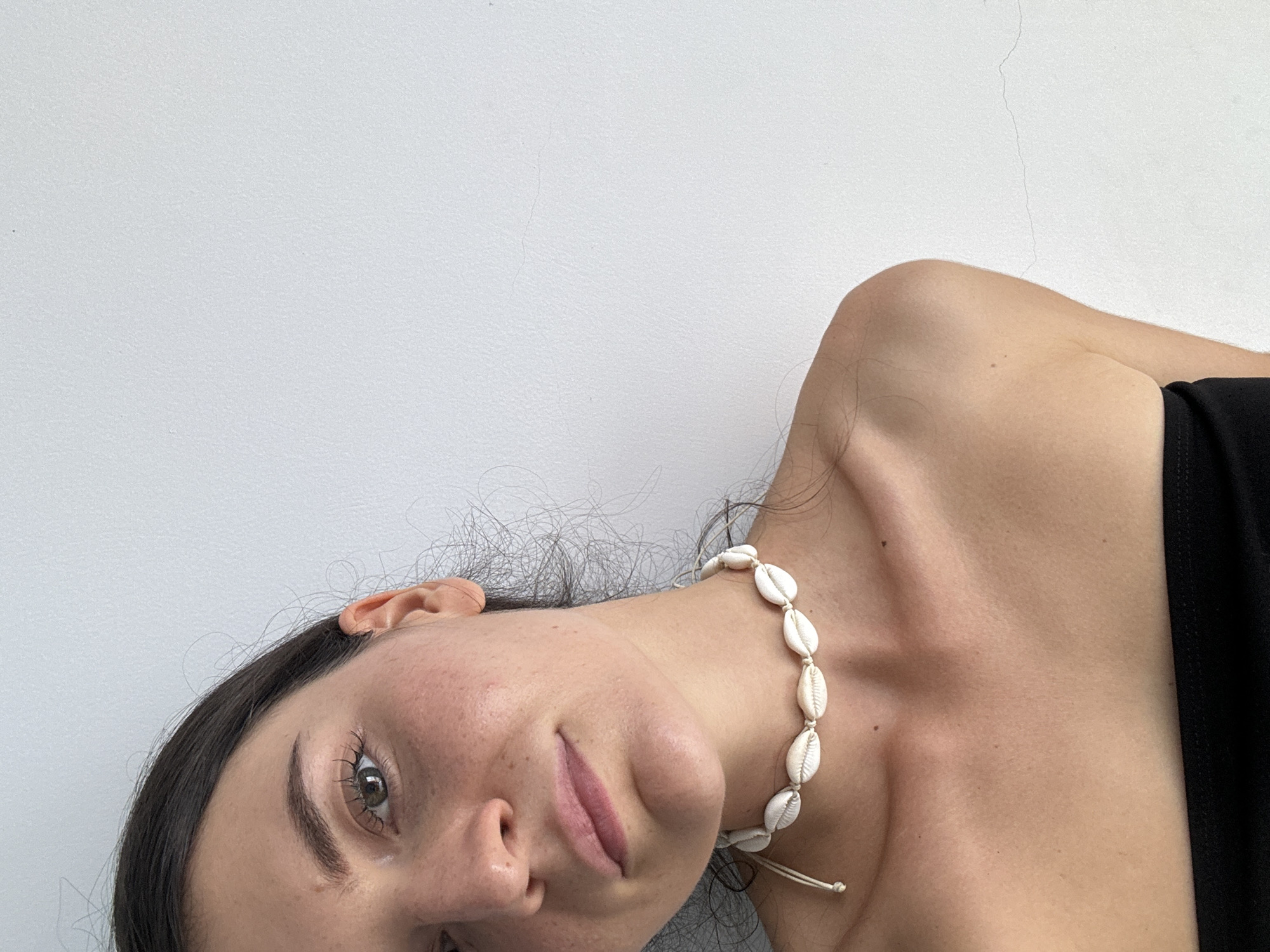A medium close-up shot depicts a woman with fair skin and dark hair, facing the camera at an angle. She wears a necklace made of small, white shells. The background is a plain, light grey wall. The location within Mũi Né, Vietnam is not determinable from the provided image.