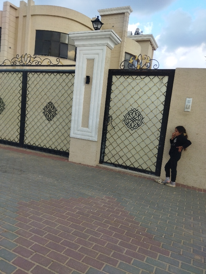 The image captures an exterior view of a modern house and its entrance gate, with a young girl standing nearby. The scene is set during the daytime under a partly cloudy sky.
The main subject is a large, two-story house with a light, possibly beige or cream, exterior. It features prominent architectural elements such as decorative pilasters and cornices. Large, dark-tinted windows are visible on the upper floor. In front of the house is an ornate metal gate, divided into two large sections, flanking a central decorative pillar. The gate itself has a diamond-grid pattern with intricate black scrollwork designs at the center of each section. A black lantern-style lamp is mounted on top of the central pillar.
To the right of the gate, a young girl, possibly between 6-10 years old, stands leaning against the house wall. She is dressed in dark clothing, possibly a tracksuit, and has her hair tied up. Her left hand is on her hip, and her right hand is raised towards her mouth, suggesting she might be eating or biting her nails, or simply resting her head. Her expression is not clearly visible. On the wall near her, there is a small white rectangular object or plaque with some unreadable text.
The ground in the foreground is paved with interlocking bricks in a pattern of alternating grey and reddish-brown sections. The overall impression is a residential setting in what appears to be a well-maintained area. Given the context, this scene is located in Khan Yunis, Palestinian Territory.