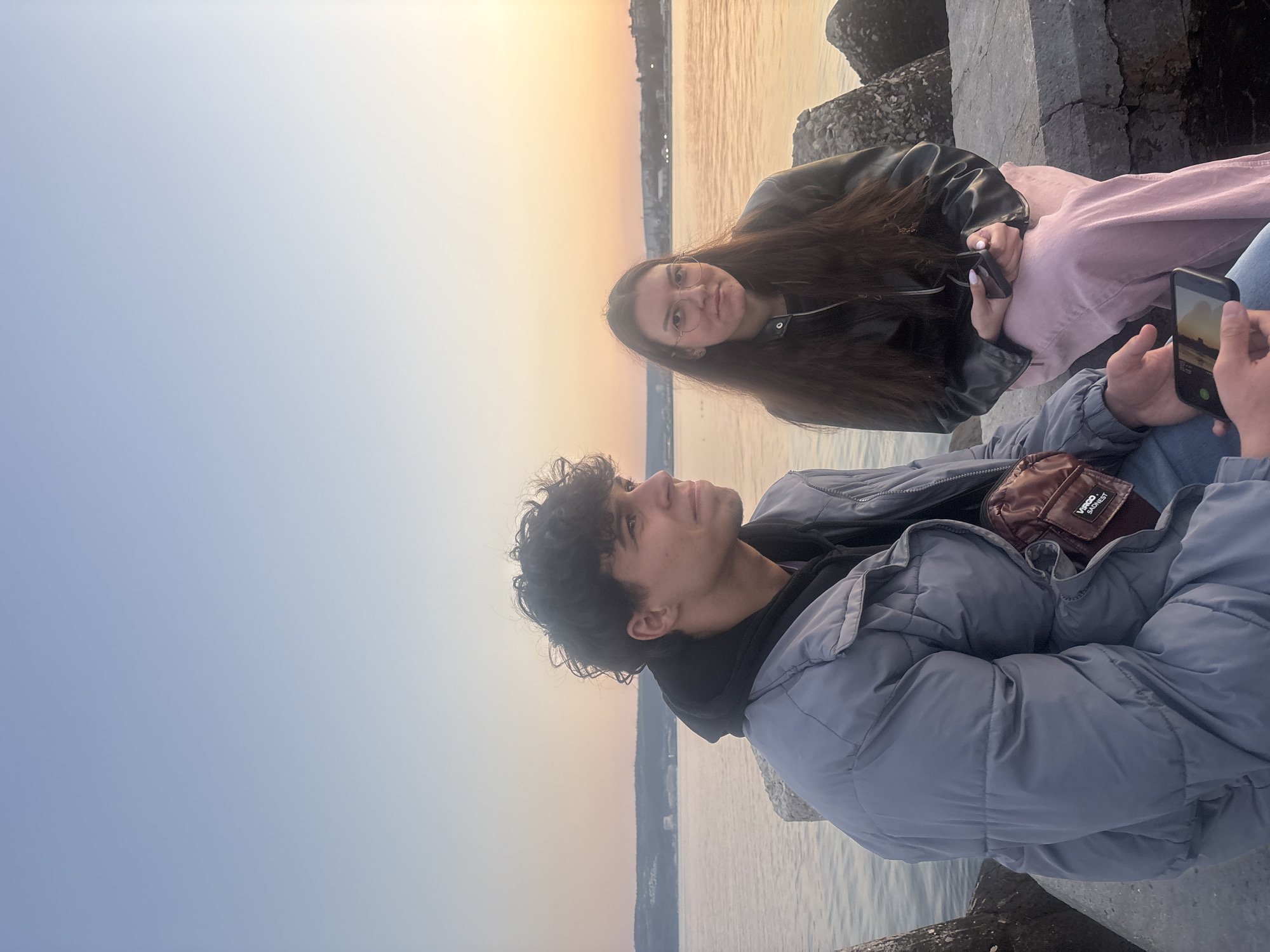 Two people are sitting on a concrete structure near a body of water, possibly a pier or breakwater. One person, on the right, is wearing a blue puffer jacket, a black hoodie, and a small brown side bag. They are holding a phone, possibly taking a picture of the sunset. The other person, on the left, is wearing a black leather jacket and pink pants. They are holding a small object in their hands. The sky in the background transitions from blue to orange, suggesting either sunrise or sunset. There are buildings visible across the water. The location is Unknown, Unknown.
