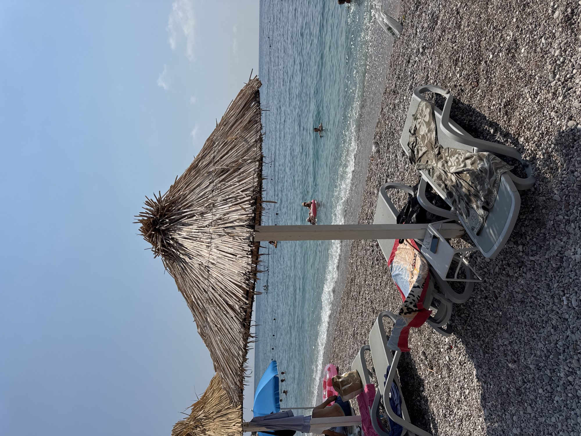 The image depicts a sunny day at a pebble beach in Mirište, Montenegro. Two thatched umbrellas provide shade over grey lounge chairs on the shore. One chair in the foreground has a towel with a camouflage pattern draped over it. A small white table sits between two chairs, with a phone on its surface. In the background, the clear blue sea is visible with several people swimming and enjoying the water. Some are using inflatable rings. The sky is bright and clear with a few wispy clouds.