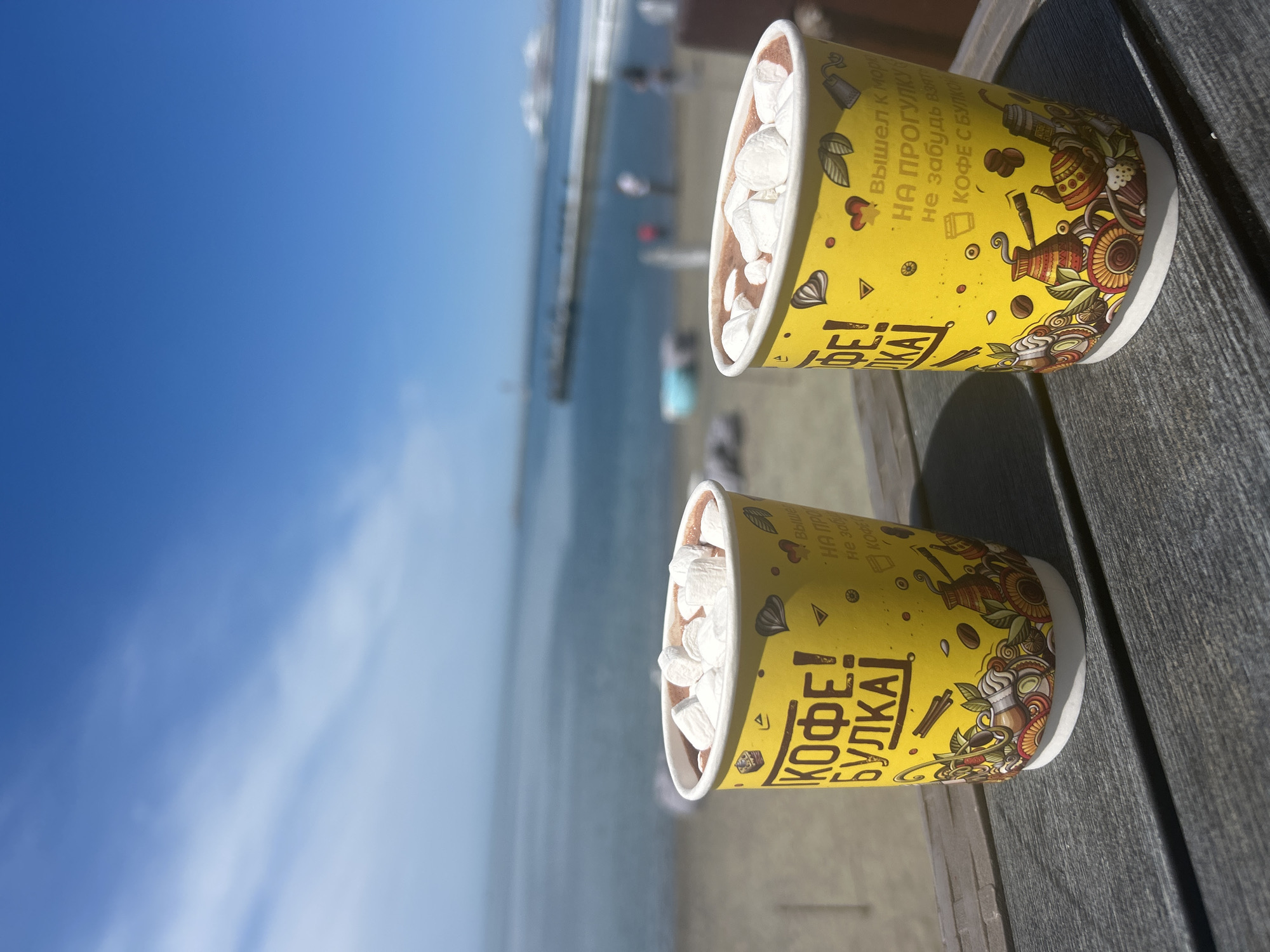 Here is a detailed description of the image:
The image features two yellow coffee cups filled with a chocolate drink and topped with marshmallows. The cups are placed on a grey wooden surface, possibly a table. The cups have Russian text printed on them.
In the background, a beach scene is visible with the ocean and a clear blue sky. There are some blurred figures on the beach, suggesting people are present. The overall scene appears to be taken outdoors in a coastal area, possibly in the city of Unknown, Unknown.