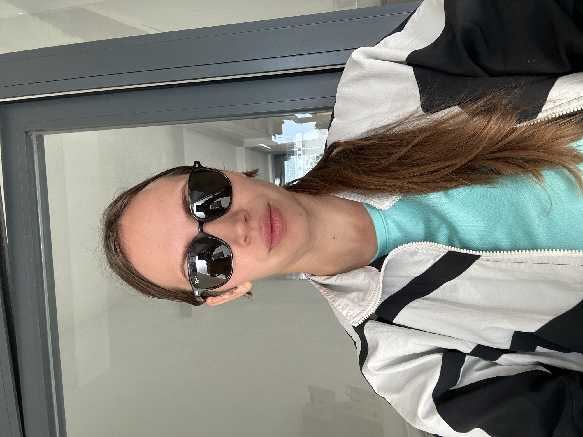 A young woman is captured in a close-up, chest-up portrait, looking directly at the camera with a relaxed, slightly smiling expression. Her long, straight brown hair is visible, flowing over her right shoulder and chest. She is wearing dark, black-framed sunglasses with reflective lenses, on which the "Ray-Ban" logo is distinctly visible.
She is dressed in a light blue collared top, layered beneath a stylish color-blocked jacket that features prominent black and white sections, possibly with a silver or grey accent, and a visible white zipper.
The background primarily consists of a large glass window or door with a dark grey or black metallic frame. Reflections in the glass show hints of an interior space with light-colored walls and possibly some distant outdoor elements. The lighting suggests it is daytime. The overall setting implies she is indoors or on a balcony next to a large window, located in Fethiye, Turkey.