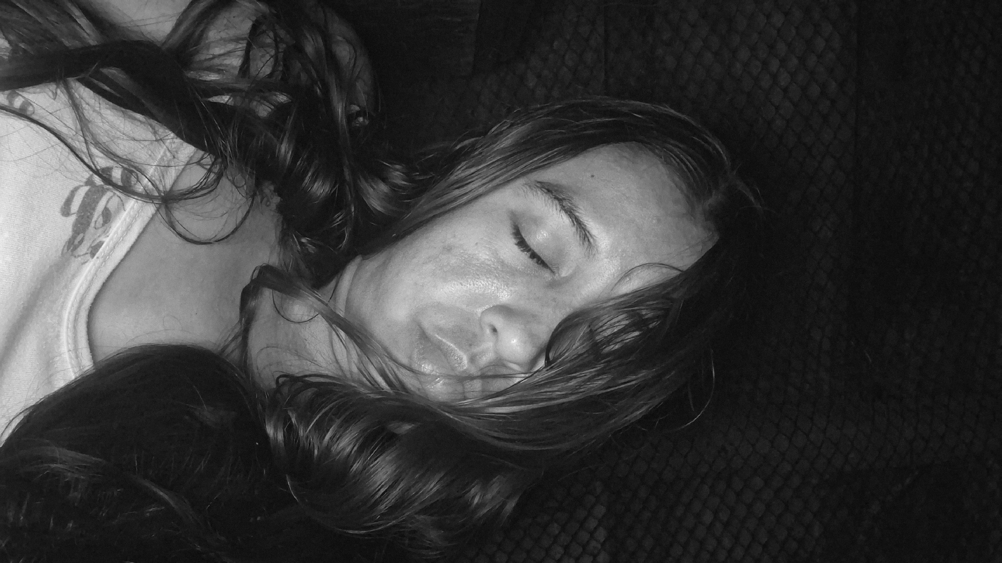 The image is a black and white, close-up, low-angle shot of a young person lying down with their eyes closed and their lips pursed as if kissing. Their hair is dark and wavy, framing their face and spilling onto their shoulder and chest. They are wearing a white tank top. The background is dark and textured, possibly a blanket or upholstery. The lighting is dim, casting shadows on the face. There are no identifiable landmarks or elements that would indicate a specific location within the city of Vallecito, Venezuela.