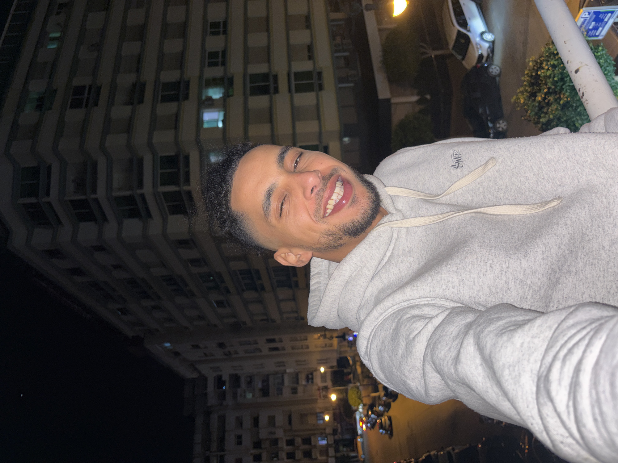 A nighttime selfie captures a smiling male in the city of Tanger, Morocco. The subject, an adult male with short dark curly hair, a beard, and a mustache, occupies the foreground, looking directly at the camera with a wide, open-mouthed smile revealing white teeth and a slight squint of his left eye. He wears a light grey, heathered hoodie with white drawstrings and a dark embroidered emblem on the left chest.
The background depicts an urban street scene under artificial illumination. Directly behind the subject are multi-story apartment buildings, light-colored with darker trim, some displaying illuminated windows with greenish or bluish light. Below, a street with parked vehicles, including a prominent white SUV on the right and darker cars on the left, is visible. Multiple streetlights cast warm yellow-orange light across the street and adjacent areas. Sparse green vegetation is present along the sidewalk. A white metal railing, exhibiting minor surface discoloration, extends horizontally across the lower right foreground.