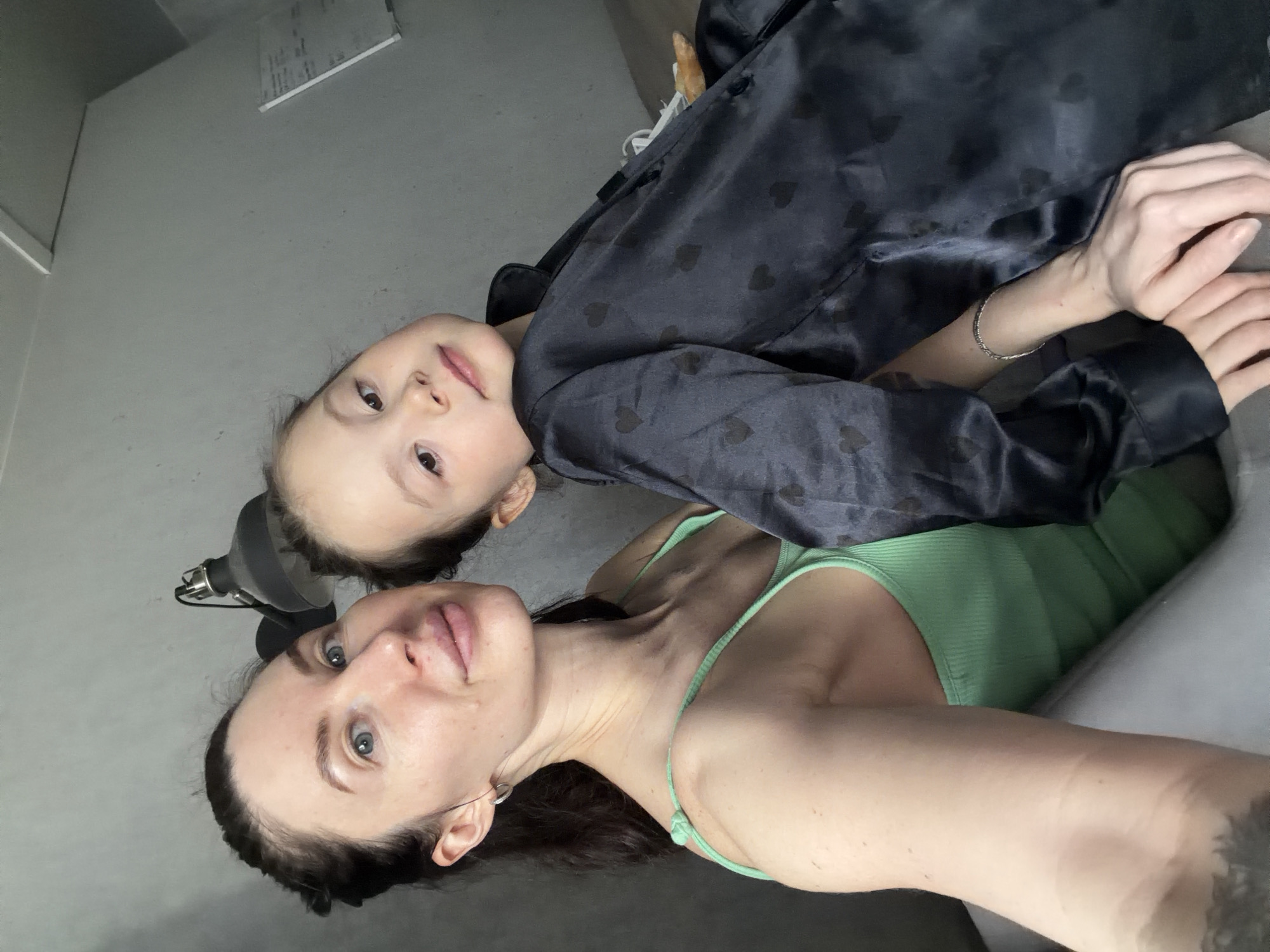 A selfie-style photograph shows a woman and a child lying down close together. The woman is wearing a light green tank top, and the child is wearing a dark-colored, patterned button-down shirt. The woman's left arm and the child's hands are visible. A lamp is visible to the left side of the frame. The location is potentially Perm, Russia, based on the user's request.