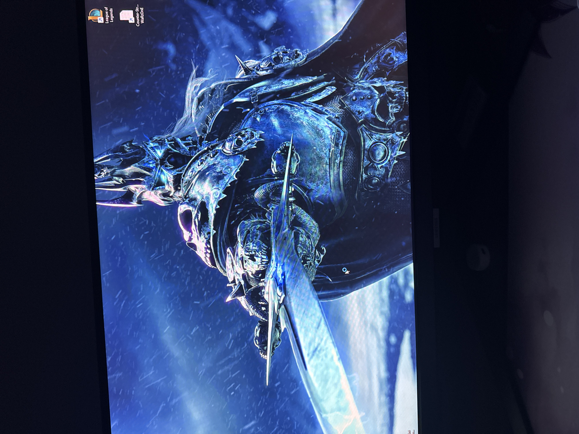 The image shows a computer screen displaying a desktop wallpaper of the Lich King Arthas Menethil from the Warcraft universe. The character is depicted wearing ornate blue and silver armor, wielding the Frostmourne sword. The background is a snowy, icy landscape. Two desktop icons are visible in the upper left corner: "League of Legends" and "Counter-Sir... WarROonE." The location is the user's desktop environment.