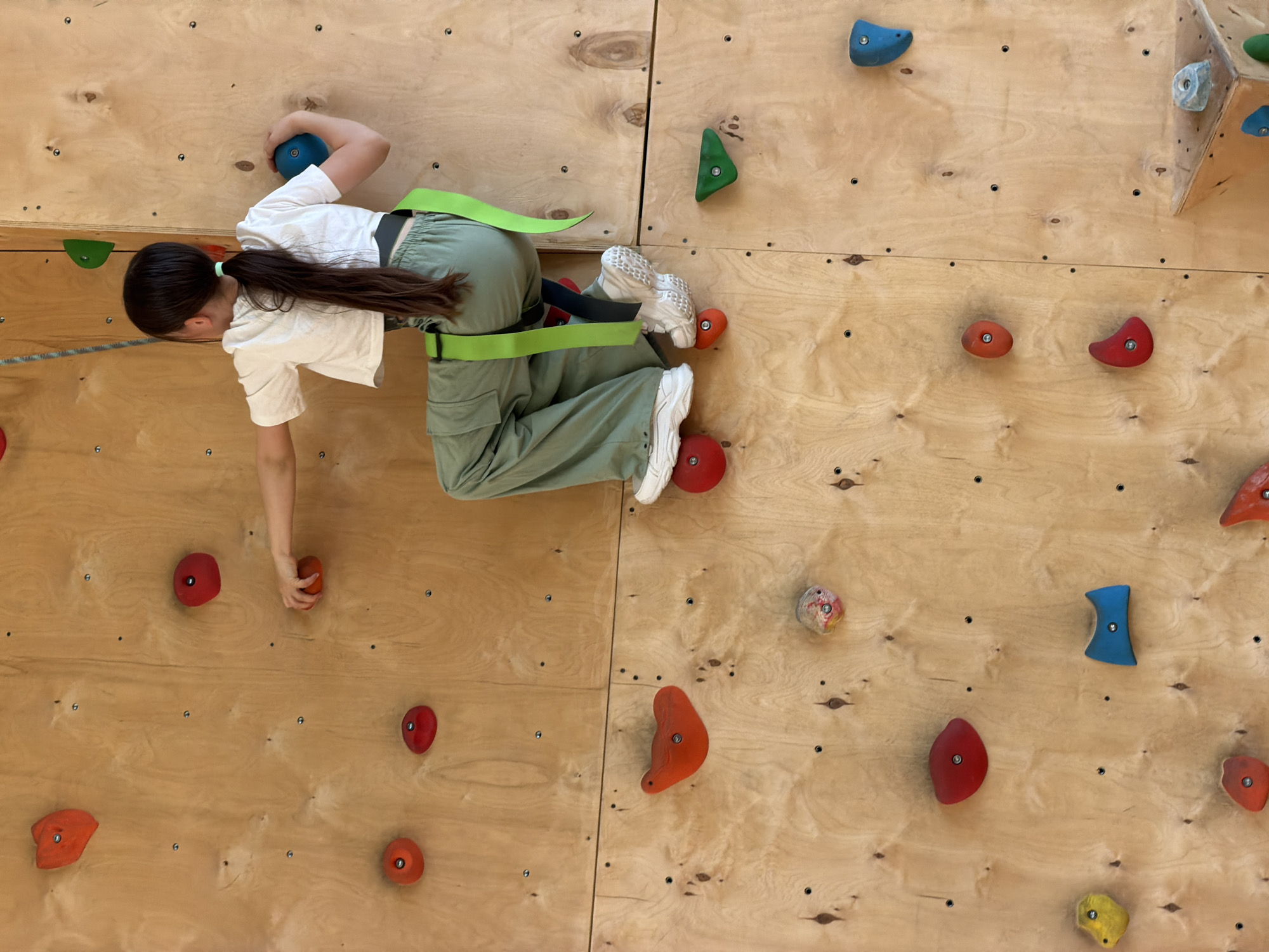A young woman with long brown hair is climbing a wooden indoor climbing wall. She wears a white short-sleeved t-shirt, green cargo pants with a green climbing harness, and white sneakers. Her right hand grips a red climbing hold while her left hand grips a blue hold higher up. Her feet are positioned on two orange holds. The climbing wall is made of light-colored wood panels with multiple colorful climbing holds attached. The scene appears to be indoors, possibly a climbing gym. It is not possible to discern a specific location in Izhevsk, Russia, from the image.