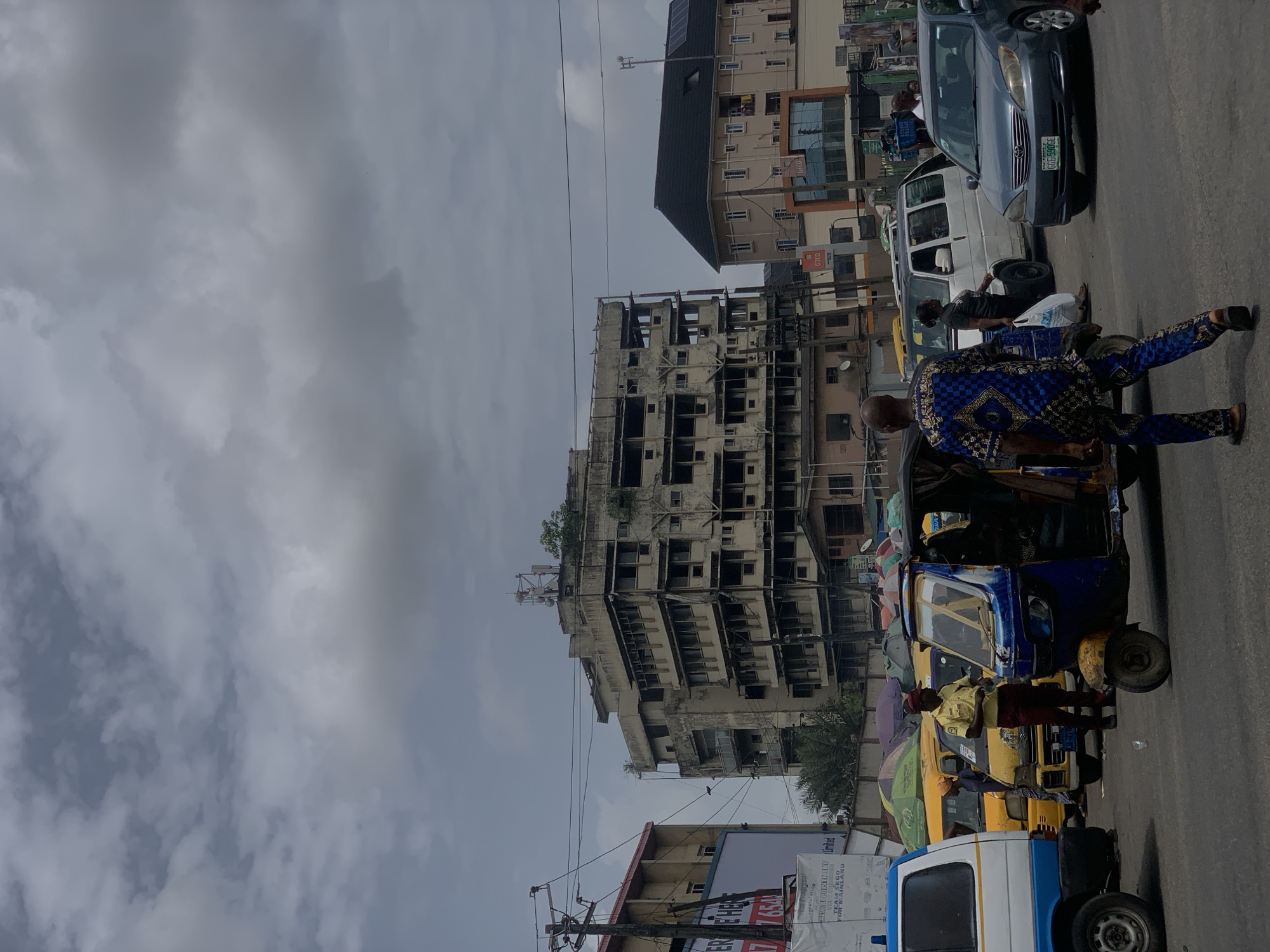 The image depicts a street scene in Alagbede, Nigeria, under a cloudy sky. A partially constructed or dilapidated multistory building dominates the background. In the foreground, several "keke" auto-rickshaws are clustered together, with passengers boarding or alighting. People are present, some dressed in traditional attire, interacting with the vehicles. Various buildings are visible, along with power lines and signage. Cars are navigating through the traffic.