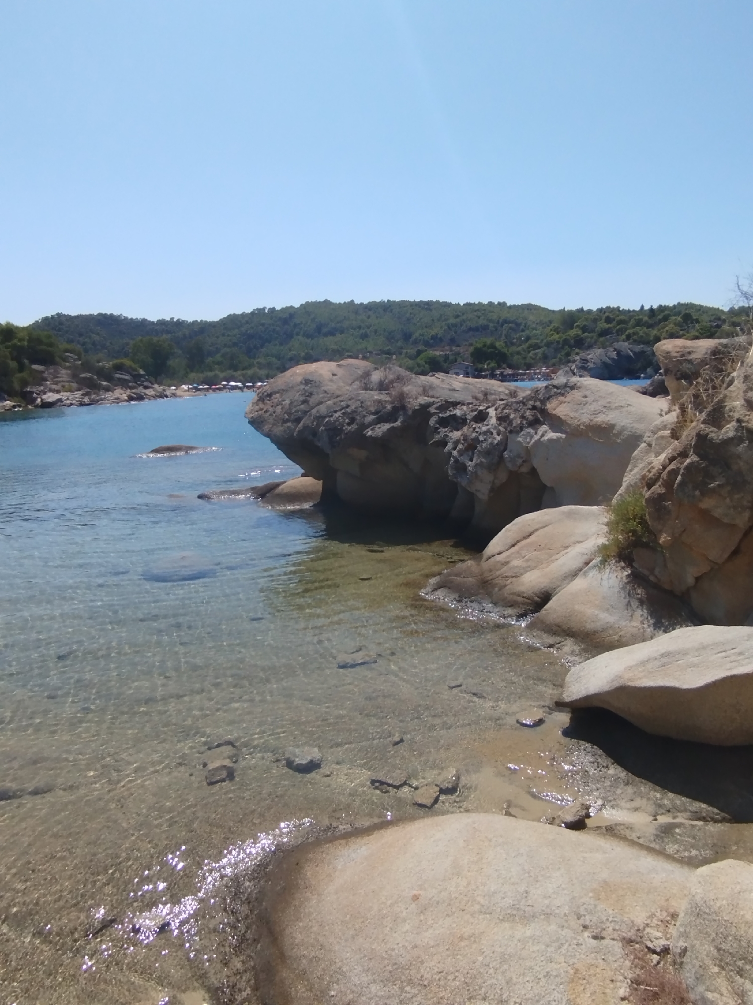 The image depicts a scenic coastal view in the Sithonia Municipal Unit, Greece. The foreground is dominated by large, weathered rock formations and a shallow, clear sea. Sunlight glints off the water, revealing the sandy seabed and submerged rocks. In the midground, the water deepens to a vibrant blue as it approaches a shoreline lined with more rocks and a stretch of beach with scattered umbrellas and what appear to be several people. The background features a lush, green, tree-covered hillside under a clear blue sky. The overall impression is one of a tranquil, sun-drenched beach day.