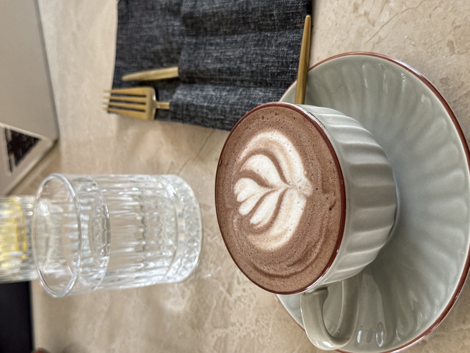 This media file captures an overhead view of a table setting in what appears to be a cafe or restaurant, located in the Ubud District, Indonesia. The central subject is a light grey ceramic cup containing a dark brown beverage, likely hot chocolate or mocha, adorned with distinct white latte art depicting a stylized heart or leaf design. A slim golden spoon is placed within the cup. The cup rests on a matching light grey saucer featuring a reddish-brown rim.
To the left of the beverage, a clear, vertically ribbed glass holds water, reflecting ambient light. Further to the upper left, a set of golden cutlery, specifically a fork and a knife, is neatly wrapped in a dark grey, textured fabric napkin. The table surface beneath these items is a light, possibly beige or cream-colored, textured material, which could be marble or stone. A blurry, light-toned object is partially visible in the top-left corner, possibly indicating a laptop or part of another table item. The scene suggests a calm and relaxed moment, perhaps for a snack or a drink. The lighting is soft and diffused, characteristic of an indoor or shaded outdoor setting during the daytime. No people are visible, and there is no discernible text within the image.