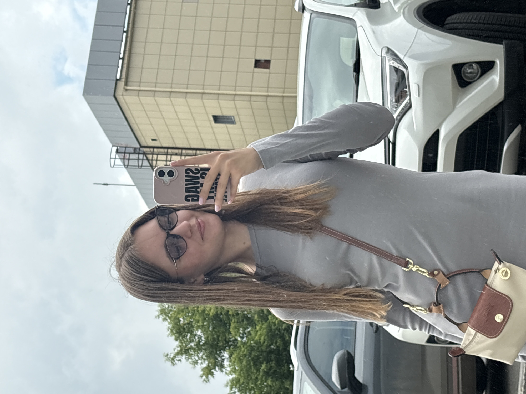 A young woman with long brown hair, wearing glasses and a grey long-sleeved dress, takes a mirror selfie. She is holding a pink phone with the word "SWAG" printed on it. A tan and brown crossbody bag hangs from her shoulder. She is standing in front of a white SUV, with a dark-colored car parked next to it. In the background, there is a building with beige tiled walls and a grey roof, and a green tree. The sky is cloudy. The scene appears to be a parking lot, possibly in an urban environment. The location is the city of Perm, Russia.