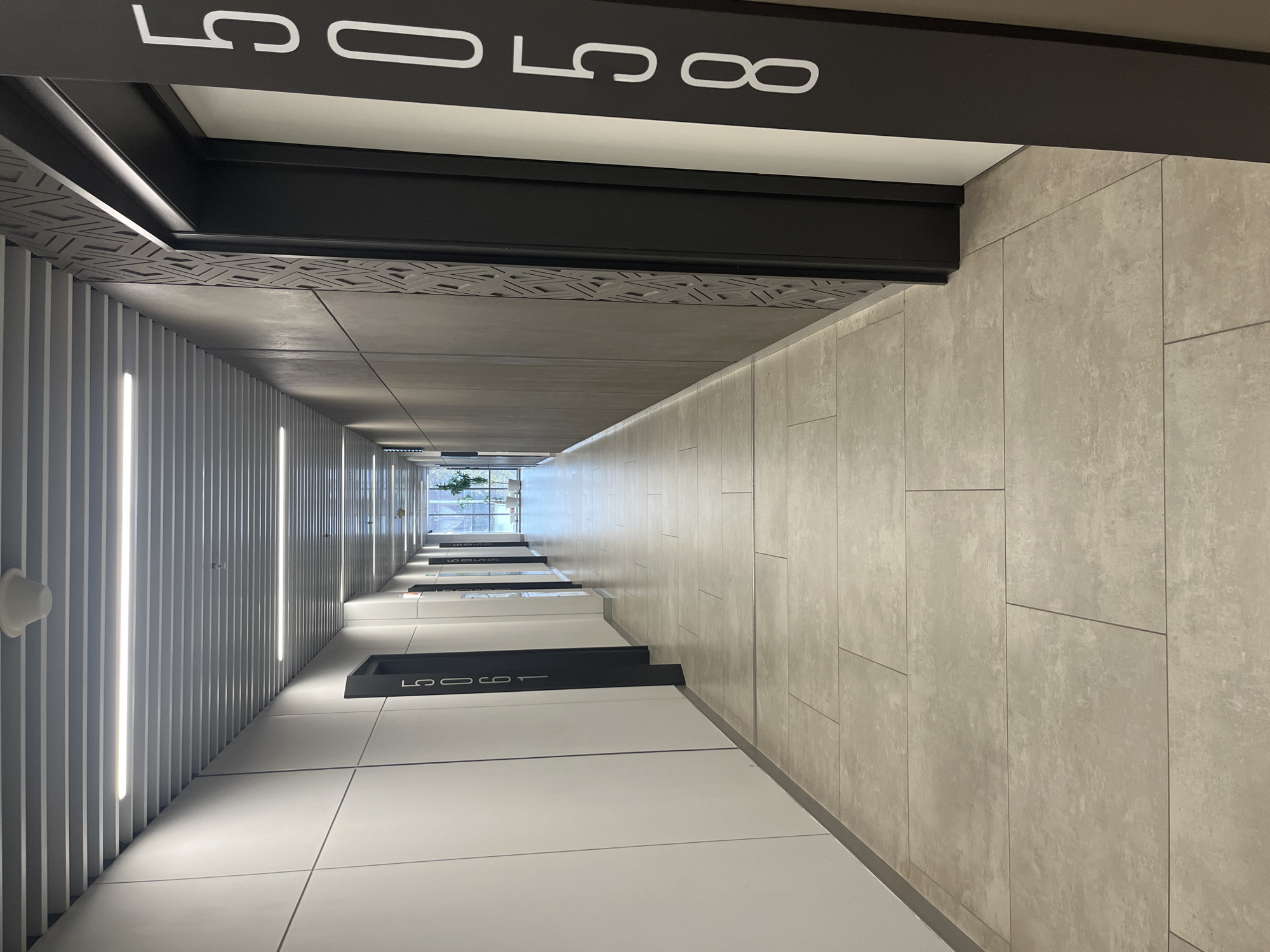 This image captures a vertical perspective looking down into a modern building's internal shaft or atrium, likely in Moscow, Russia. No people are visible.
The scene showcases contemporary architectural design elements, characterized by clean lines and a monochromatic palette of greys, blacks, and whites. On the right side of the frame, a wall is clad in large, light grey, textured tiles, possibly resembling concrete. The left side features a wall composed of vertical slats or panels, with integrated linear LED light strips running vertically, casting a bright glow and creating a rhythmic pattern down the shaft.
The top of the image shows the underside of an upper floor or balcony, a prominent black beam with white, stylized numbers: "50 58". Further down the shaft, similar black structures with white numbering are visible on lower levels, sequentially marking "50 57", "50 56", and "50 55". These numbers likely denote apartment or room numbers on different floors. A decorative geometric pattern can be seen on a dark-colored soffit or beam near the top left.
At the very bottom of the shaft, a window or opening reveals natural light and a hint of green foliage outside, suggesting an exterior view or an internal courtyard. The overall impression is one of modern, minimalist design, emphasizing verticality and light. No specific activity or event is taking place; the image is a static architectural study of the space.