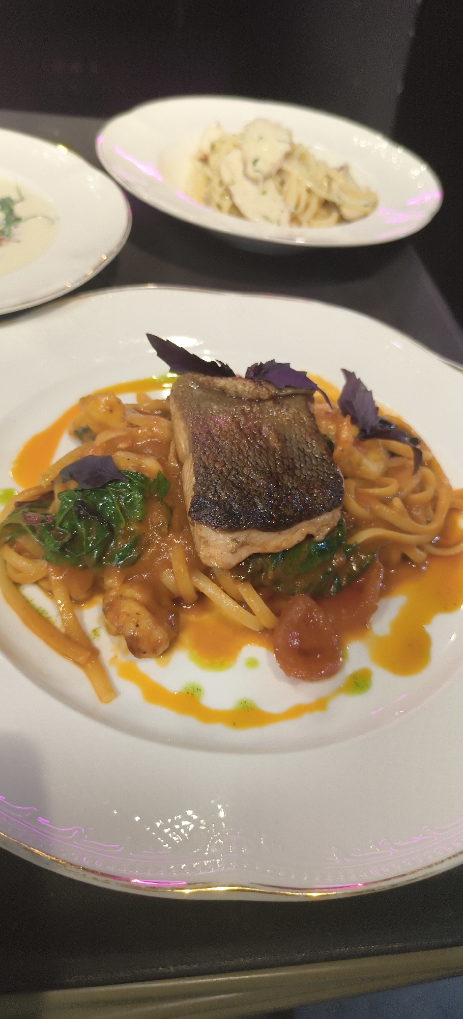 A close-up shot captures a plate of food, likely a meal, in a dining setting. The primary dish on the plate consists of pasta with a reddish-orange sauce, topped with a piece of fish, possibly salmon, with crispy skin. Green vegetables and purple basil leaves garnish the dish. The plate itself is white with a gold rim. In the blurred background, two other plates are visible: one containing a cream-colored soup, and another with pasta in a white sauce. The image was taken indoors on a dark surface, likely a table. Given the location context of Yerevan, Armenia, this could be a restaurant specializing in European or fusion cuisine. No people are visible in the shot.