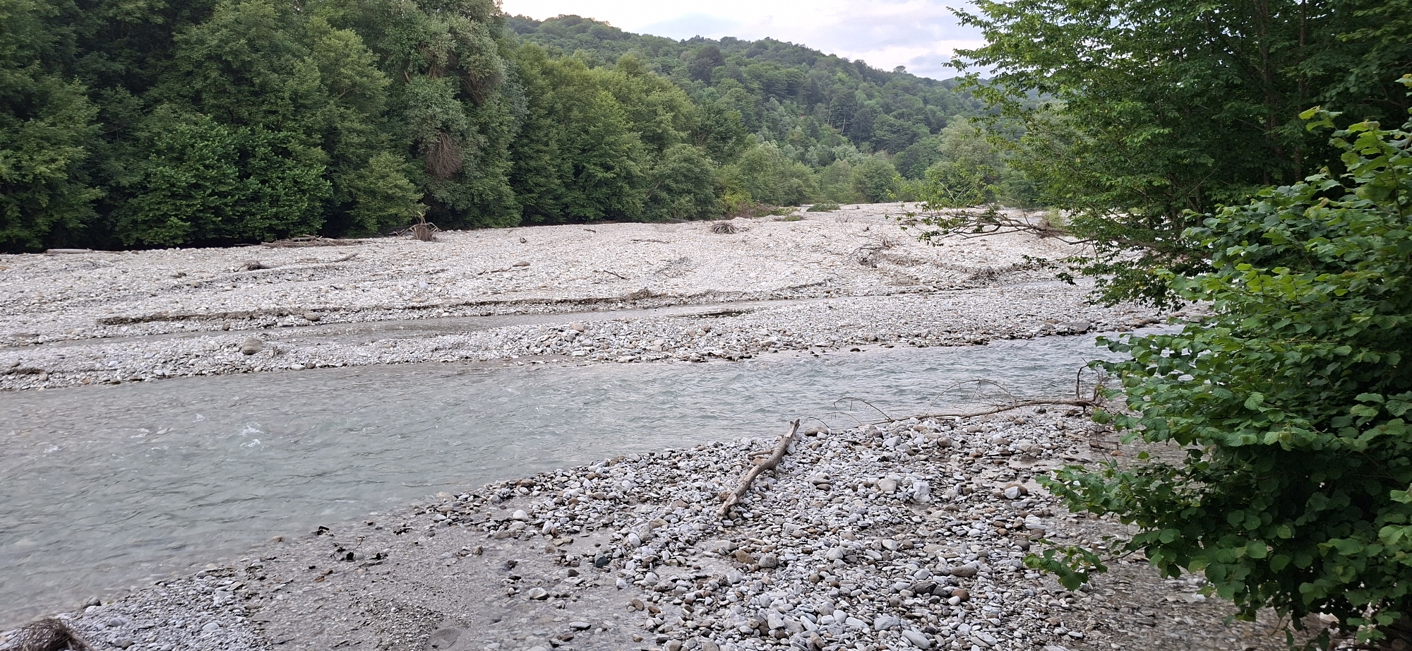 A wide, shallow river flows through a rocky, sparsely vegetated riverbed. The water is a murky grey-blue and appears to be moving slowly. The riverbed is composed of smooth, rounded grey and white pebbles of varying sizes, with some larger rocks and driftwood scattered throughout. On either side of the river, dense green trees cover the slopes of rolling hills. The sky above is overcast and pale grey. The scene depicts a natural landscape that appears to be located in the city of Serzhen-Yurt, Russia.