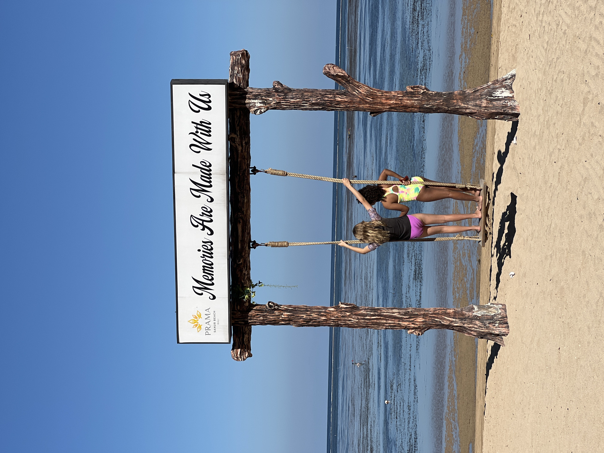 Two children stand on a rope swing in front of a sign that reads "Memories Are Made With Us" with the logo "PRAMA Sanur Beach Bali" on it. The swing is made of rope and wooden planks, suspended between two faux-wooden support beams holding the sign. The children, one with dark hair and a yellow and pink swimsuit and the other with blonde hair and a black top and pink shorts, are standing on the swing facing forward towards the water. The background consists of a calm ocean and a clear blue sky. The setting appears to be a beach in Sanur Beach, Bali, part of the city of Denpasar, Indonesia.