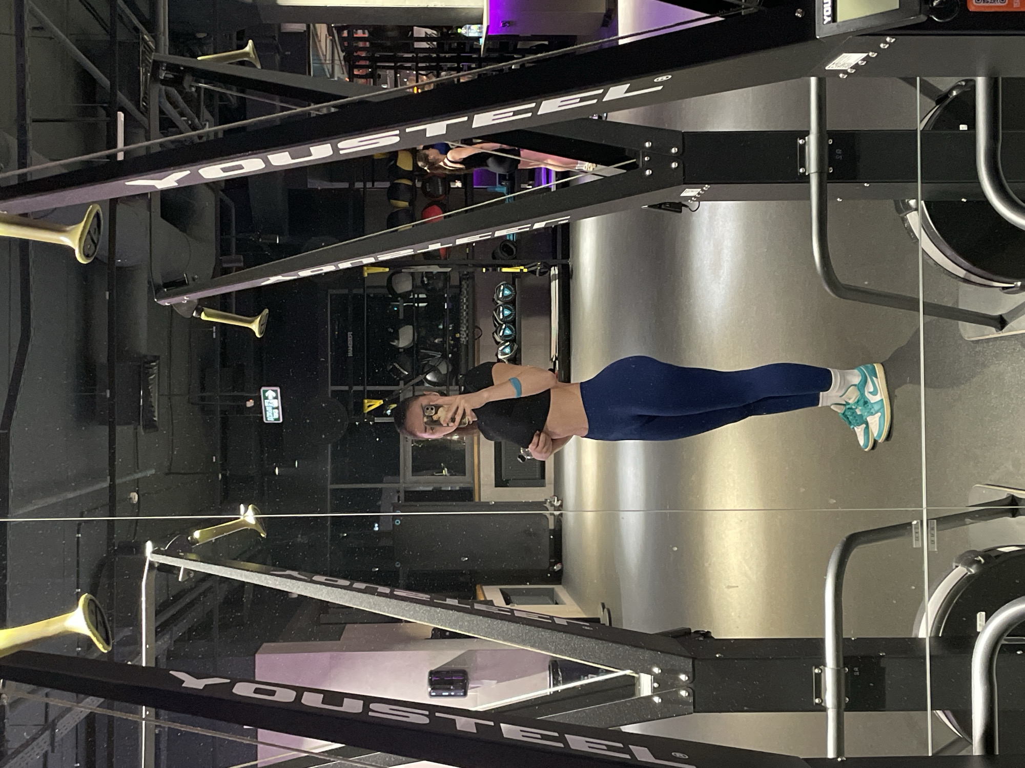 A young woman with brown hair pulled back is standing in a gym, taking a mirror selfie. She is wearing a black crop top, navy blue leggings, and teal and white sneakers. Her left arm is extended, holding a smartphone, and her right hand is on her abdomen. She is positioned in front of a large black and silver gym machine with the word "YOUSTEEL" printed vertically on it. The gym environment is visible in the background, with other gym equipment, mirrors, and lighting. The floor is dark and reflective. The overall lighting suggests an indoor setting, possibly at night or with artificial lighting. The location of the gym is not specified in the image.