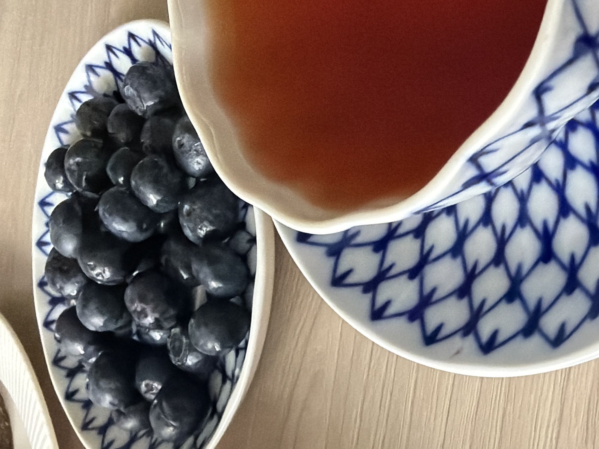 A top-down shot depicts a cup of tea and a dish of blueberries, likely on a wooden table. The cup and saucer are white with a blue lattice pattern, reminiscent of the Lomonosov porcelain factory's style, a significant cultural element in Saint Petersburg, Russia. The blueberries are piled high on a small, oval dish also decorated with a blue lattice pattern. The tea is a warm, amber color. The arrangement suggests a breakfast or snack setting. The image conveys a sense of calmness and simple pleasures. The absence of additional cues prevents further localization within Saint Petersburg.