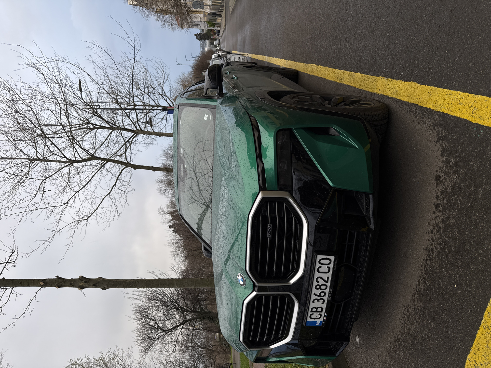 The image shows a green BMW XM parked on a street in Burgas, Bulgaria. The vehicle is parked close to a yellow line marking the edge of the roadway. The license plate reads "CB 3682 CO." It appears to be a cloudy day, and there are bare trees visible in the background. Other cars are parked further down the street.