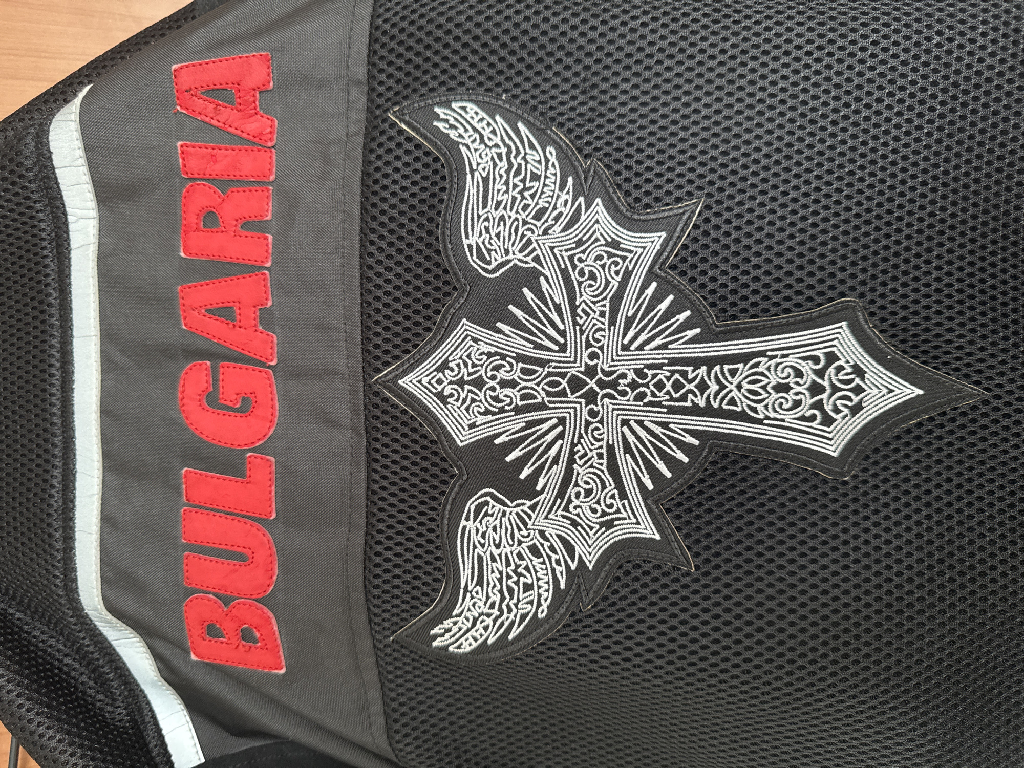 The image shows a close-up of a black mesh fabric, possibly part of a jacket or bag. On the left side, there's a vertical panel with the word "BULGARIA" embroidered in red capital letters. Next to it, there's a reflective white stripe. On the right side of the image, a black patch featuring a stylized cross with wing-like extensions on either side is visible. The cross design is outlined in white embroidery. While the image contains the word "BULGARIA", there is no evidence to indicate that the image was taken in the city of Plovdiv, Bulgaria, or any other specific location.