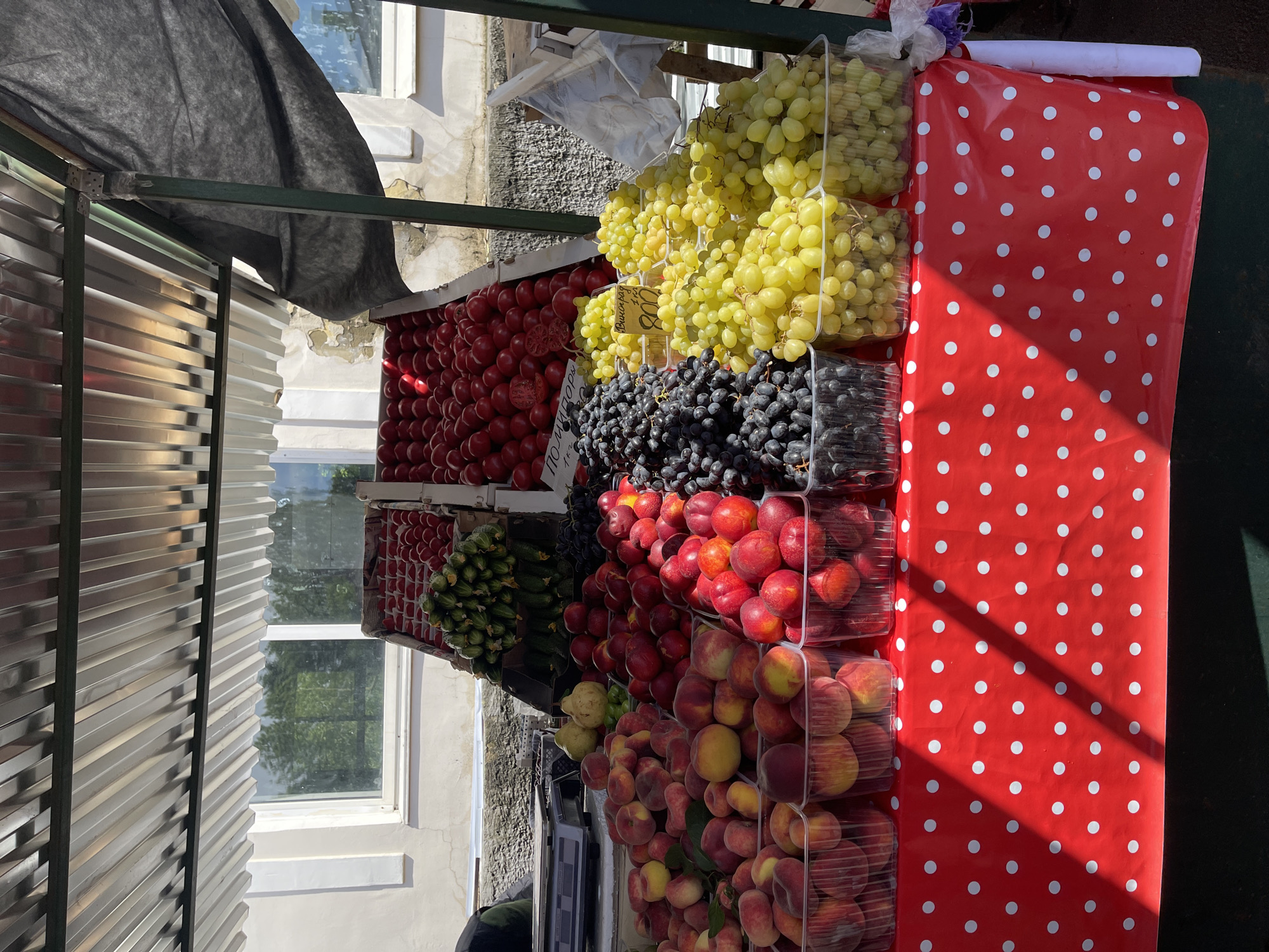 The image shows a fruit stand in an unidentified city, with rows of various fruits displayed for sale. The fruits include red tomatoes, yellow grapes, dark blue grapes, and peaches in plastic containers. A red tablecloth with white polka dots covers part of the stand. A window is visible in the background, and the stand is covered by a dark fabric shade.
