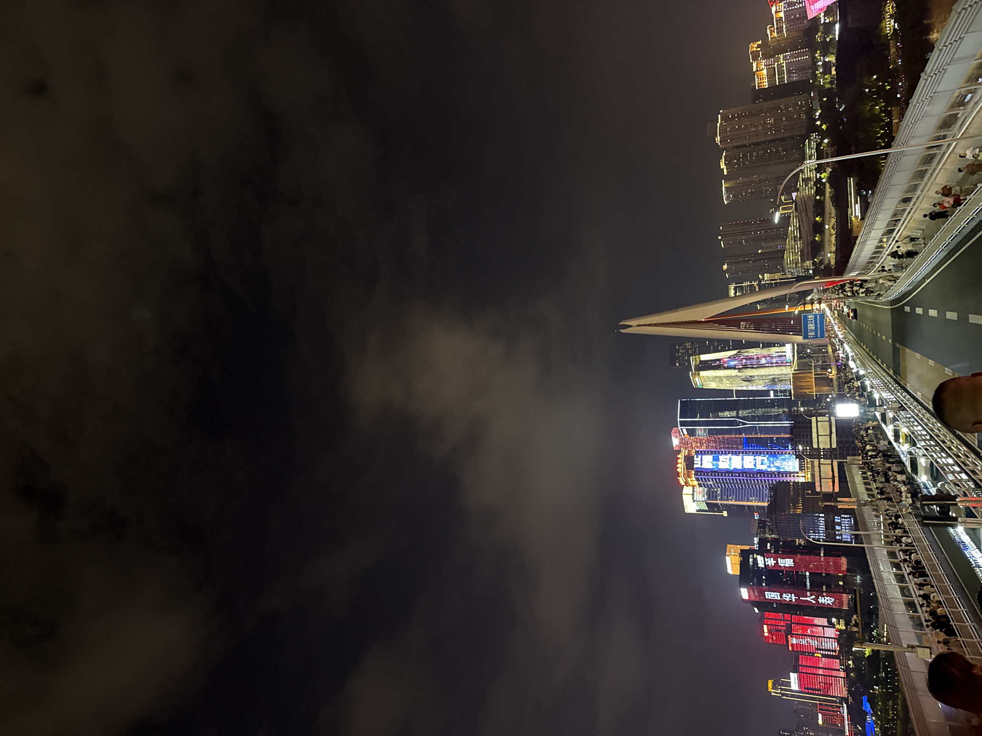 The image captures a vibrant nighttime scene of the Yuzhong District in Chongqing, China, likely from a bridge overlooking the cityscape. Tall buildings adorned with colorful illuminated advertisements and lights dominate the skyline. A prominent cable-stayed bridge with a distinctive white pylon is visible, with vehicles on the road and pedestrians walking along the dedicated pedestrian walkway. The sky is dark and partly cloudy, with the city lights reflecting off the clouds. The overall impression is of a bustling urban environment at night, illuminated by a spectacular display of artificial lights.