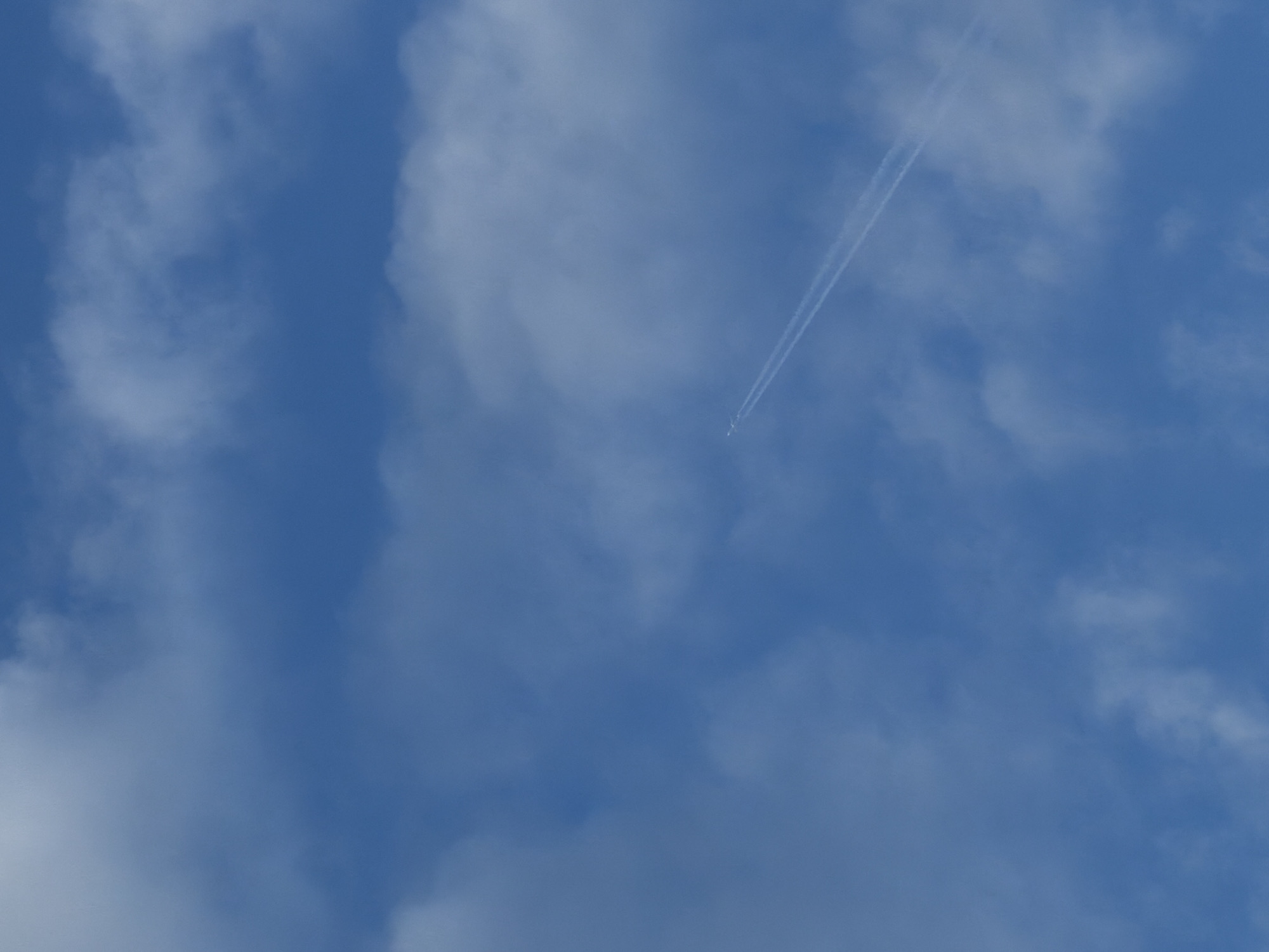 The image depicts a blue sky with scattered white clouds. A contrail, possibly from an aircraft, is visible as a pair of white lines diagonally crossing the upper right portion of the frame. The city of Chaikovsky, Russia, is not identifiable from this image.