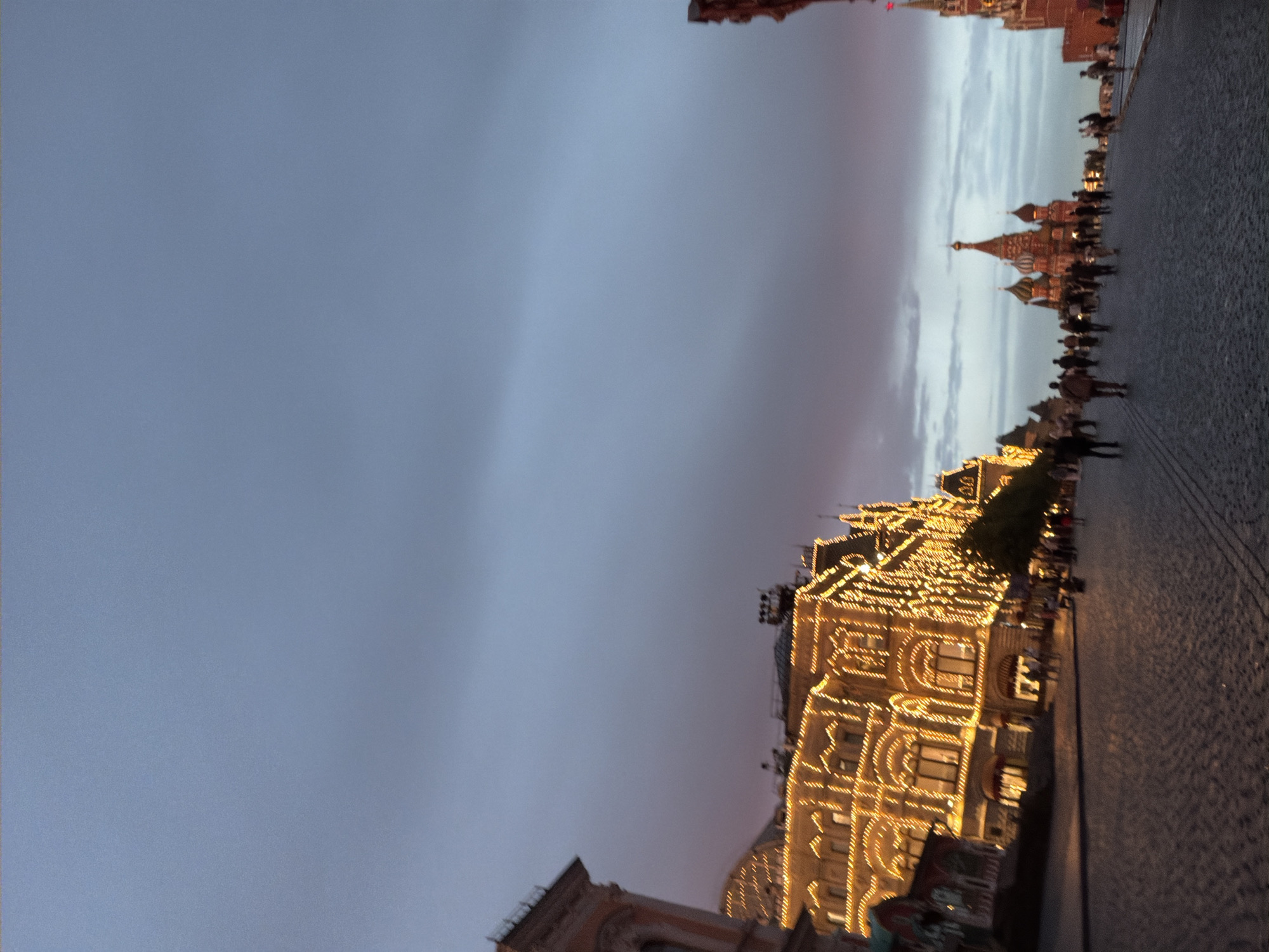 The image depicts a bustling scene in Moscow, Russia, likely on Red Square, during twilight or early evening. The sky is a soft gradient of pale blue and muted pink, hinting at the transition from day to night. A prominent, ornate building, adorned with a multitude of warm, glowing lights, dominates the right side of the frame. Its architecture suggests a grand historical structure, possibly a department store or a government building.
In front of this illuminated building, a cobblestone pathway is visible, lined with numerous people walking in various directions. The crowd appears to be a mix of tourists and locals enjoying the evening ambiance. Further in the background, the iconic onion domes of Saint Basil's Cathedral, also illuminated, pierce the dusky sky, confirming the Moscow location. The overall atmosphere is one of lively urban activity and a celebration of architectural beauty enhanced by evening lights. The weather appears calm, with no strong indications of rain or wind. The scene evokes a sense of wonder and appreciation for the historical and illuminated cityscape of Moscow at dusk. No visible text is present in the image.