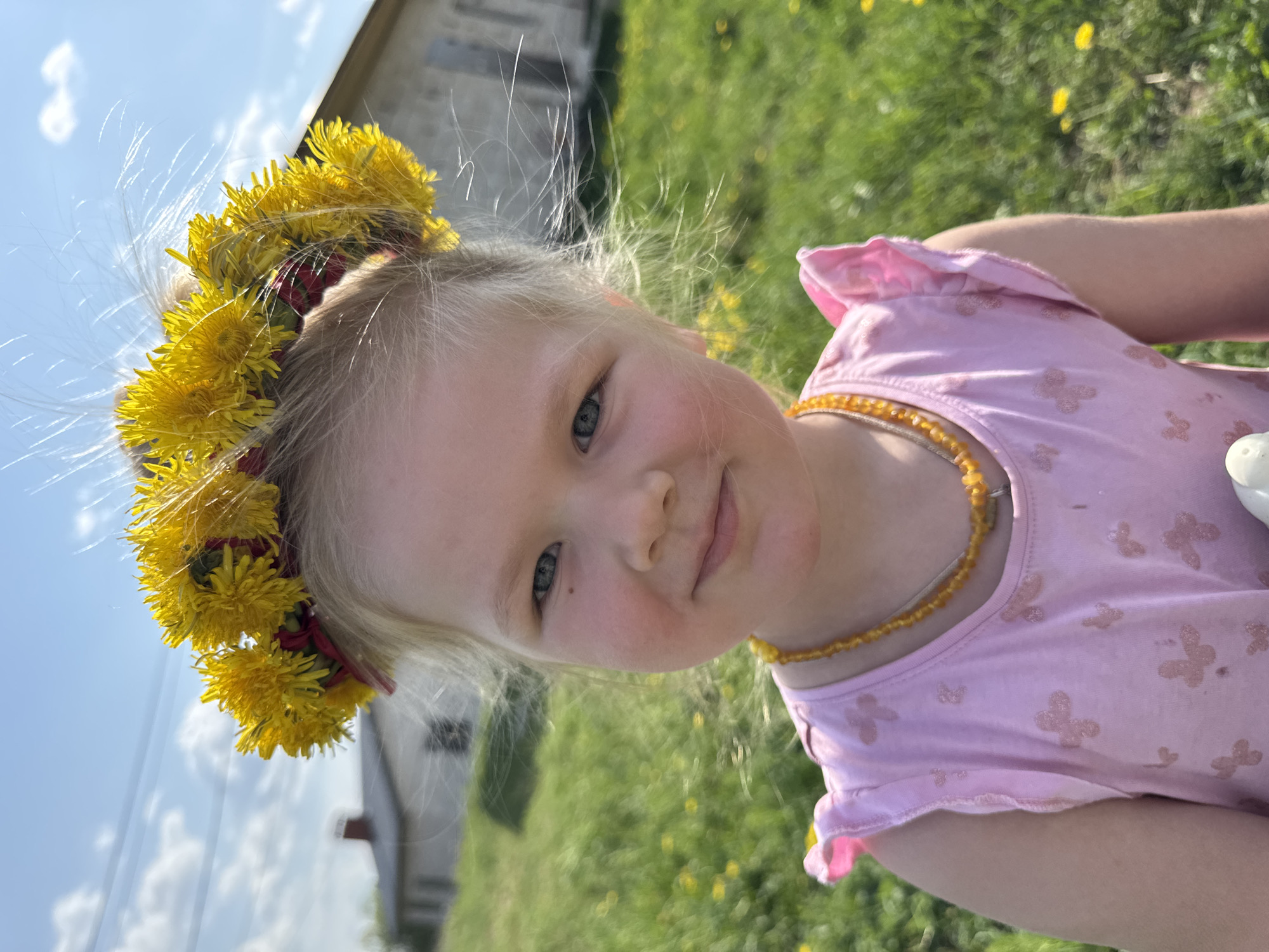 A young girl, likely between 4 and 6 years old, is centered in the image. She wears a crown of yellow flowers and a pink shirt with a necklace. The background is a grassy field dotted with dandelions, a building is visible in the background. The location is Ферма, Russia.