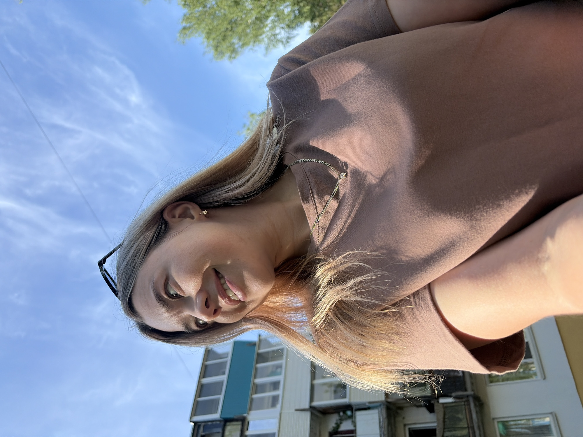 A woman with blonde hair is posing for a selfie. She is wearing a brown t-shirt and a delicate gold necklace with a small charm. Sunglasses are perched on her head. Behind her is a building in Chaikovsky, Russia, under a blue sky with a few clouds. Some greenery is visible in the upper part of the frame. The building appears to be residential, with visible windows and a teal-colored section.