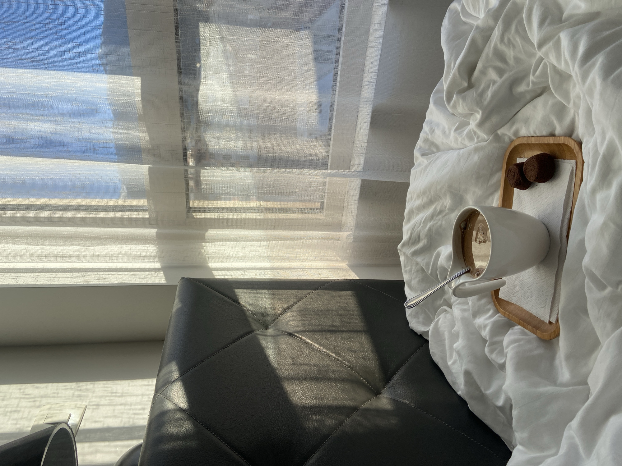 An eye-level, indoor shot shows a black leather bench or ottoman, a tray on top of white bedding, and a window with a white sheer curtain.
The leather bench/ottoman takes up the bottom left of the photo. Light streams through the window onto the bench. The bench is quilted with diamonds and thick stitches.
On top of the bench is a pile of white bedding, possibly a blanket or comforter. On top of the bedding is a small wooden tray with a white mug containing coffee or hot chocolate. A metal spoon is resting in the mug. The tray also has two dark brown chocolate cookies and a small white napkin.
Behind the bench/ottoman and bedding is a window covered with a sheer white curtain. Bright light is filtering through the window.
The location is unknown, though the prompt mentions the city of Budva, Montenegro.