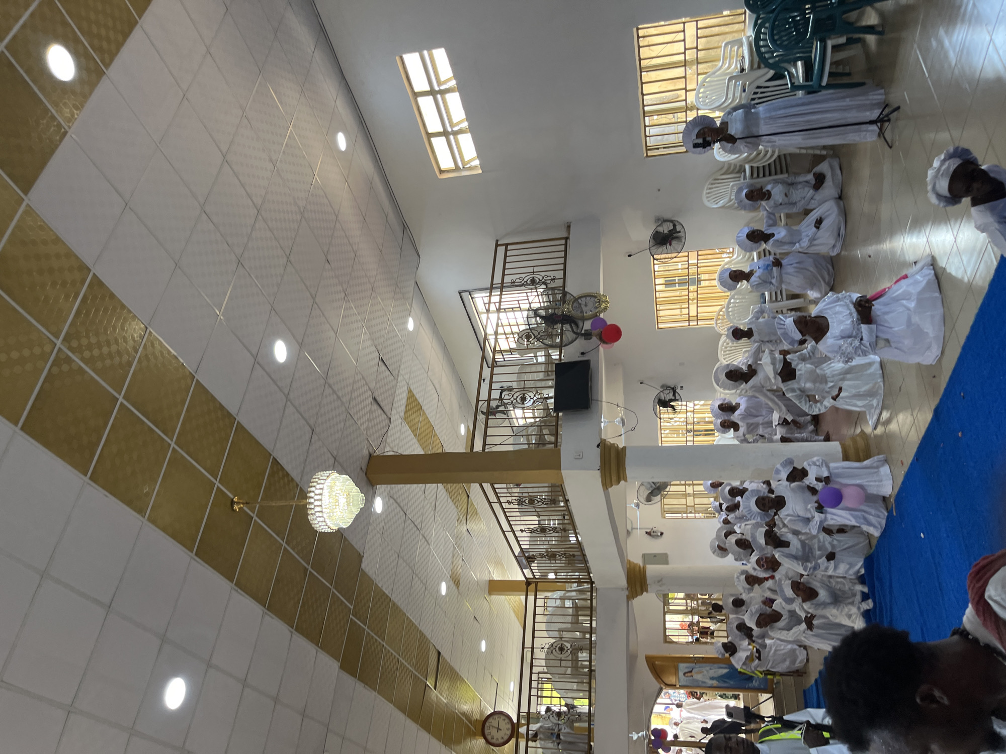The image displays an indoor gathering within a spacious hall or church. The ceiling is composed of white and gold textured tiles, featuring a prominent diagonal band of gold. It is equipped with multiple recessed lights and a central ornate chandelier. The walls are white, complemented by gold-painted columns and an upper-level mezzanine with decorative gold metal railings. Several rectangular windows with grids are visible, admitting natural light.
A large group of individuals, predominantly women and children, are present, all uniformly dressed in white robes or dresses and white head coverings. Many are depicted kneeling or sitting on the tiled floor, a portion of which is covered by a blue mat in the foreground. A woman in white is kneeling, holding one purple and one pink balloon. To the right, another woman in white stands, operating a smartphone mounted on a black tripod, presumably recording the event. Behind her, stacks of white and green plastic chairs are visible.
On the upper mezzanine, several individuals in white attire are seated on white plastic chairs. A large red-rimmed wall clock is mounted on the upper left wall, and an ornate gold wall clock hangs near the mezzanine railing on the right. A black flat-screen television is mounted on the wall below the mezzanine. Multiple black wall-mounted oscillating fans are affixed throughout the space. Through an open doorway in the background, additional individuals in white are visible, alongside a framed picture of the Virgin Mary. In the foreground, the back of a man's head is visible, and another man wearing a white shirt with a lime green vest stands to the left.
