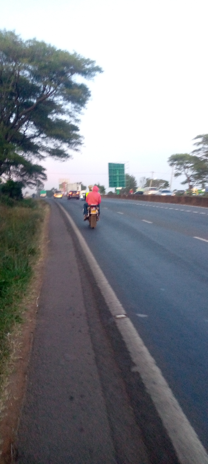 A daytime, slightly blurry shot shows a two-lane highway with a motorcycle carrying two individuals, one wearing a red hoodie. The motorcycle is centered in its lane, moving away from the camera. Additional vehicles are visible further down the road. The highway is bordered by green grass on one side, and a large green highway sign is seen in the background. Tall trees are visible on both sides of the highway, blending into the slightly overcast sky.