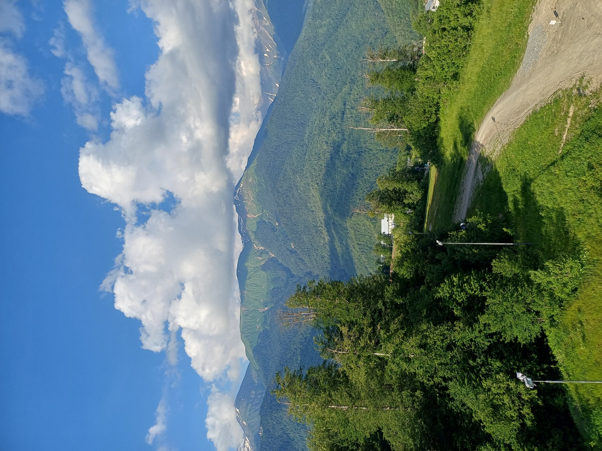 This is an outdoor, eye-level, wide shot of a mountain landscape in Esto-Sadok, Russia. The scene depicts a steep, tree-covered mountain range under a bright blue sky with scattered white clouds. A dirt path winds its way up the grassy slope on the right side of the image, casting shadows from the trees onto the grass. There are a few scattered bare trees, and some white buildings are visible in the distance nestled amongst the trees. The sun appears to be shining, suggesting it is daytime. The overall impression is one of a serene and natural mountain setting, possibly during the summer months. There is no visible text in the image.